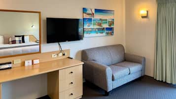 Habitación de hotel con cama, escritorio con cajones, televisor de pantalla plana, sofá gris, cuadro colgado en la pared y ventana con cortinas.
