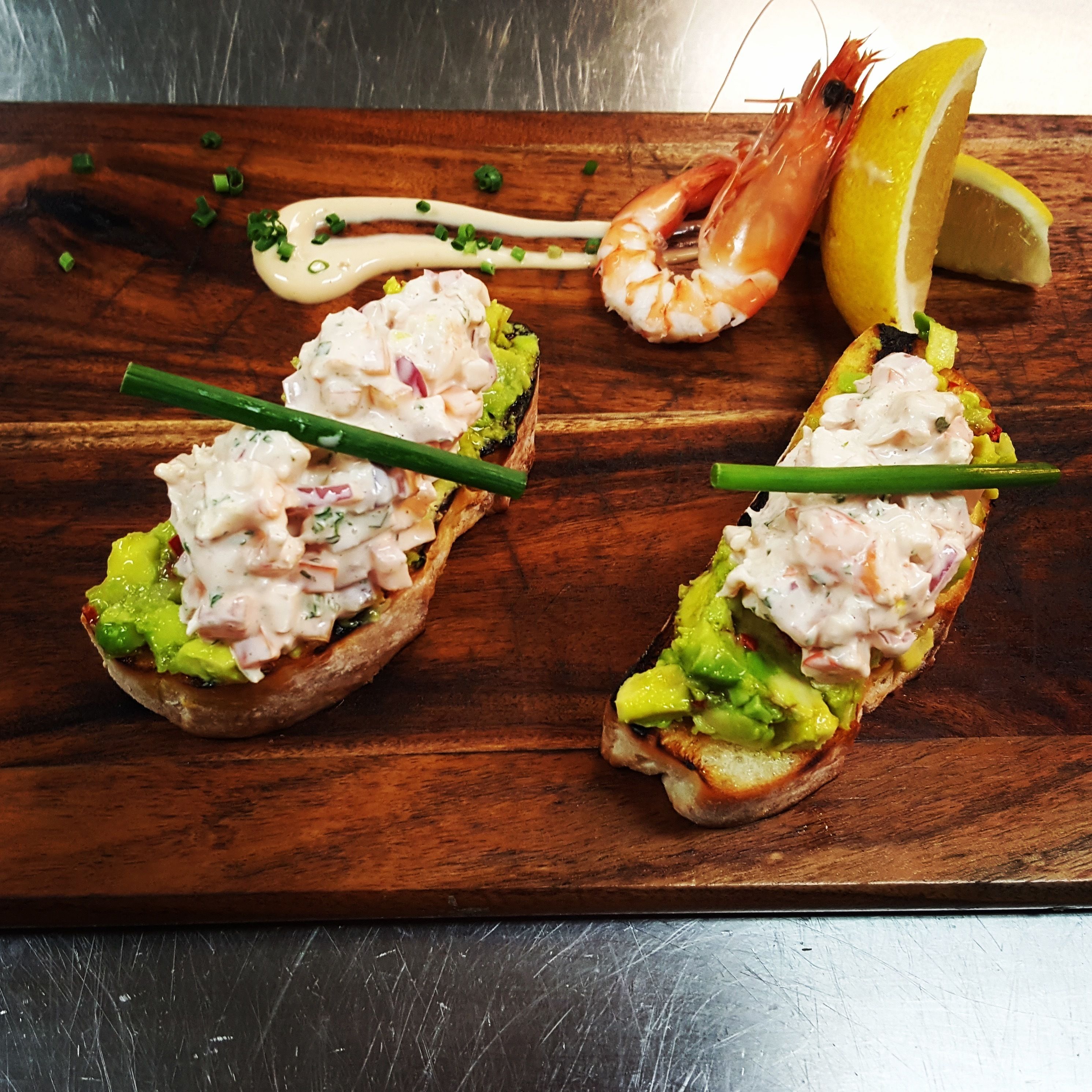Two open-faced sandwiches with avocado and a creamy topping, served on a wooden board with a lemon wedge and a shrimp.