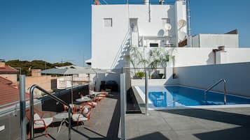 A rooftop pool area with a clear blue pool, outdoor seating, and a white building with satellite dishes.