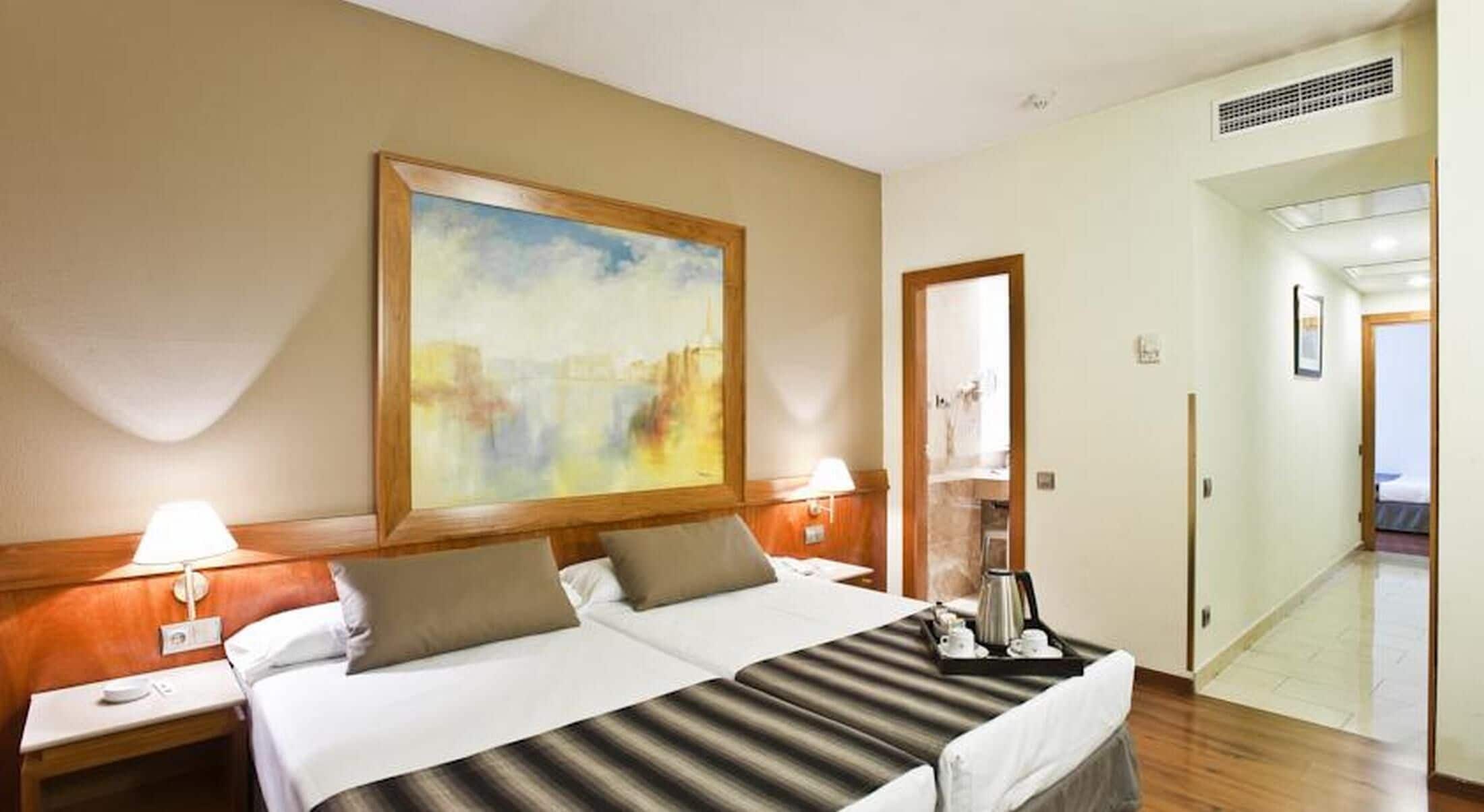 A hotel room with a bed, two bedside tables, a painting, a coffee maker, and a view of a hallway.