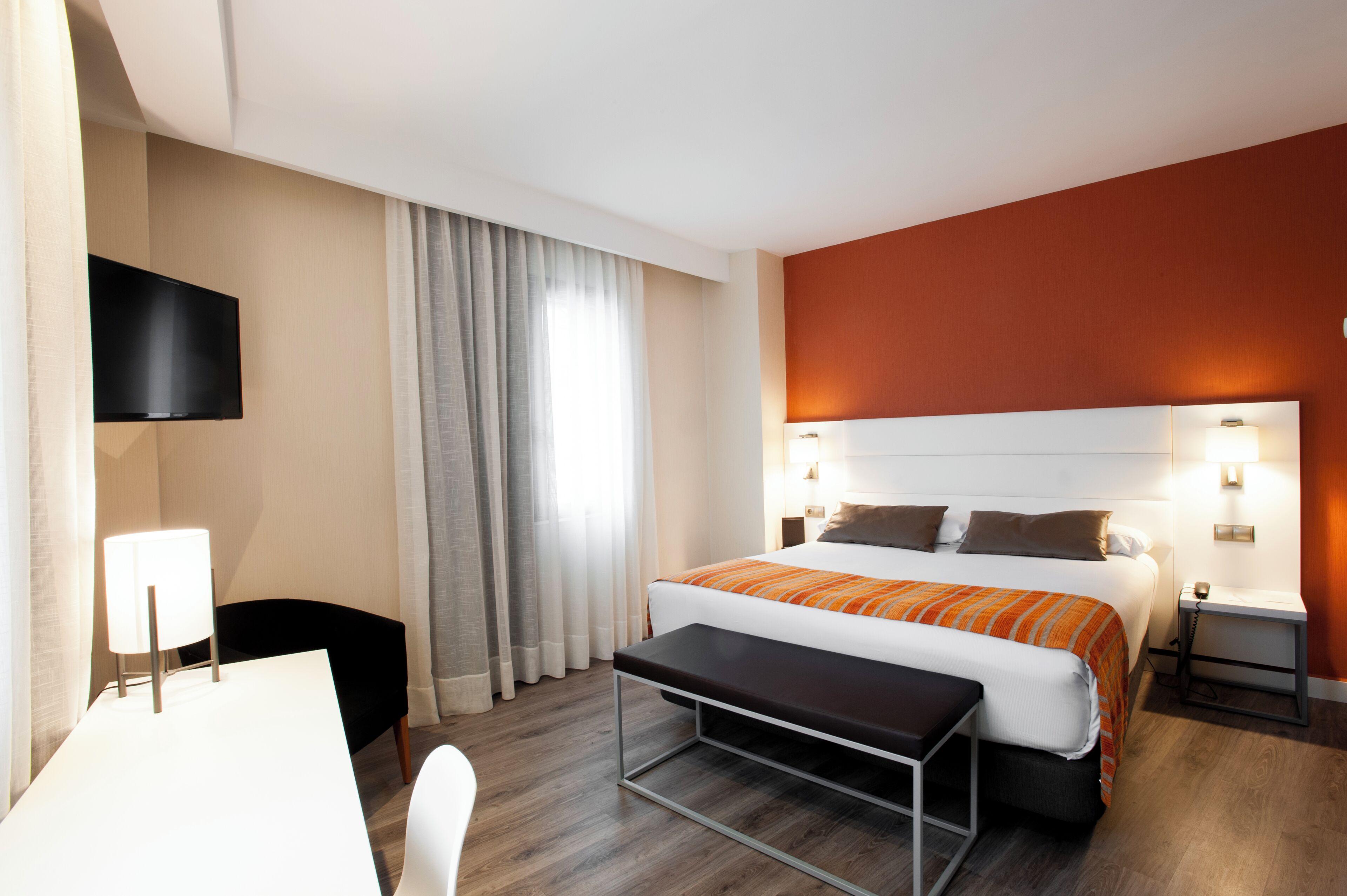 A hotel room with a bed, bedside tables, a desk, a chair, a TV, and curtains.
