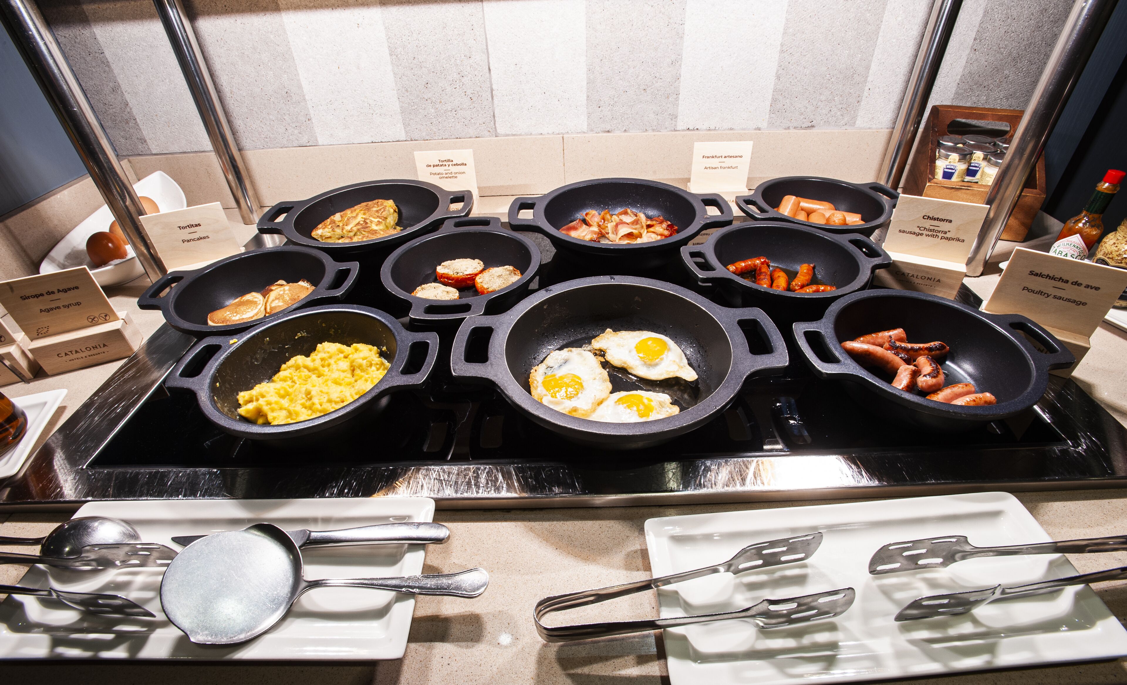 Un bufé con diversas opciones de comida, entre ellas huevos revueltos, salchichas y tortillas.