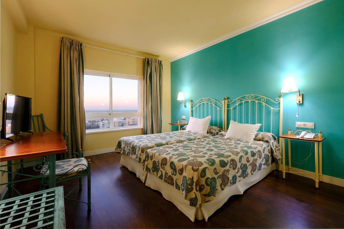 Een hotelkamer met een bed, een bureau, een stoel, een televisie, een raam met gordijnen en uitzicht op de stad.