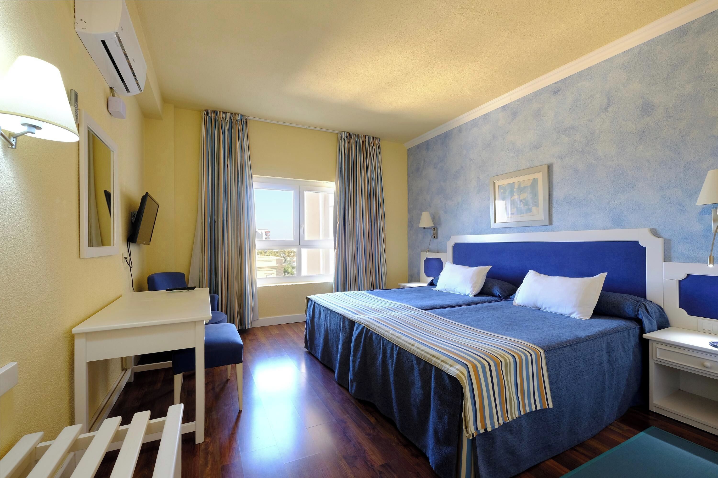 Een hotelkamer met een bed, nachtkastjes, een bureau, een stoel, een raam met gordijnen en een airconditioner.