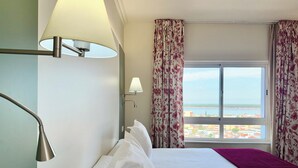 A hotel room with two beds, a window with curtains, a lamp, and a view of the beach.