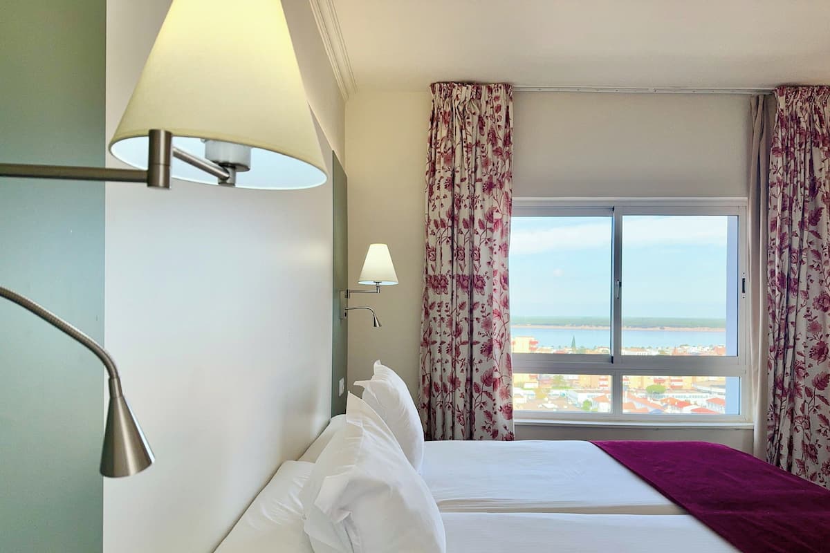 Een hotelkamer met twee bedden, een raam met gordijnen, een lamp en uitzicht op het strand.