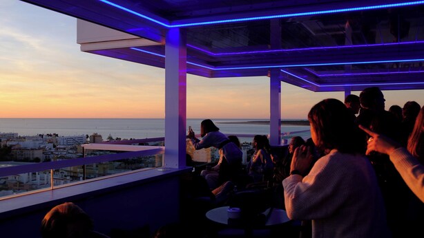 A rooftop bar with a view of the city and the sea at sunset.