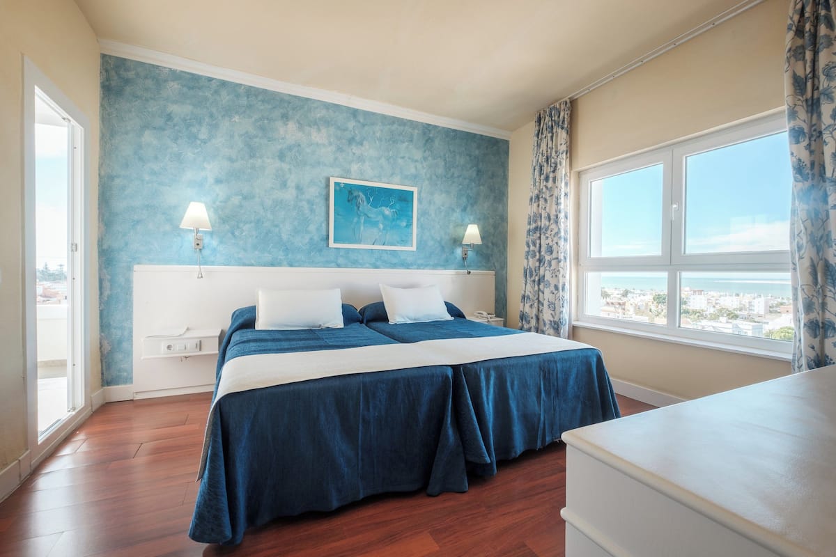 Een tweepersoonsbed met een blauwe spreideken, een nachtkastje, een lamp en een raam met uitzicht op zee.