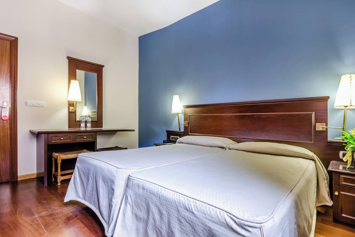 A hotel room with a large bed, wooden headboard, bedside tables with lamps, a desk with a chair, and a mirror.