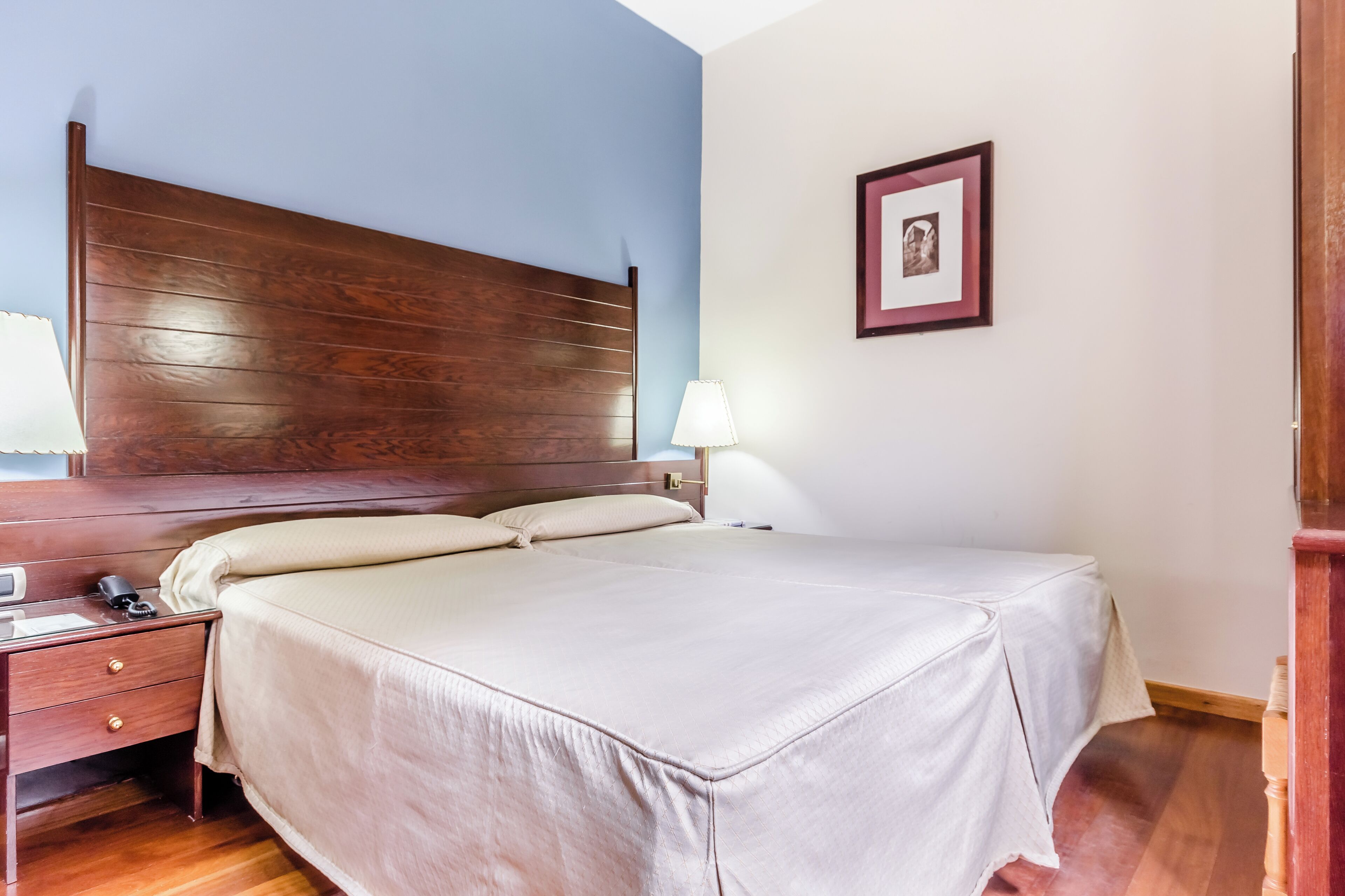 Una cama bien hecha con cabecera de madera, una mesita de noche con lámpara y un cuadro enmarcado en la pared.