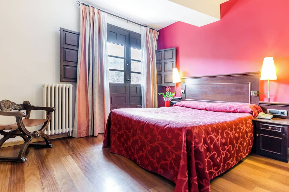 A hotel room with a large bed, a wooden nightstand, a desk, and a radiator.