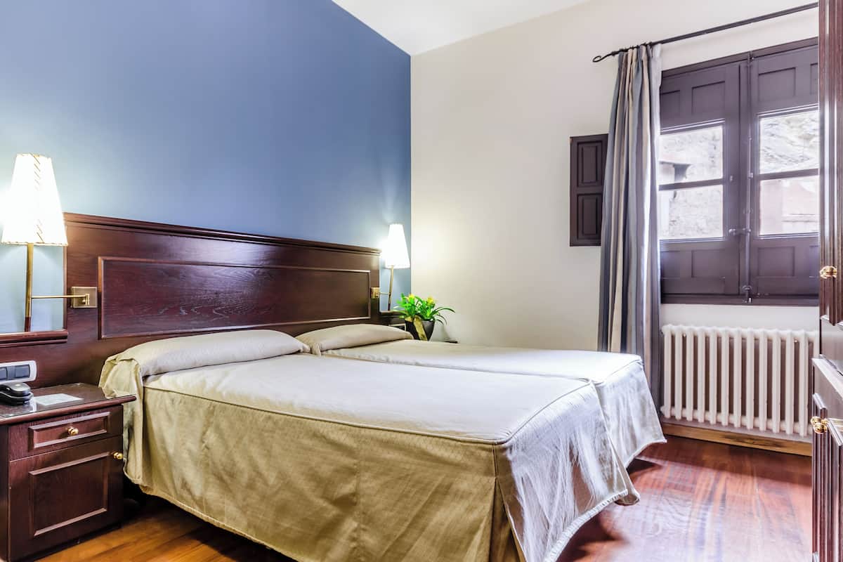 A double bed with a wooden headboard, bedside tables with lamps, a potted plant, and a window with curtains.