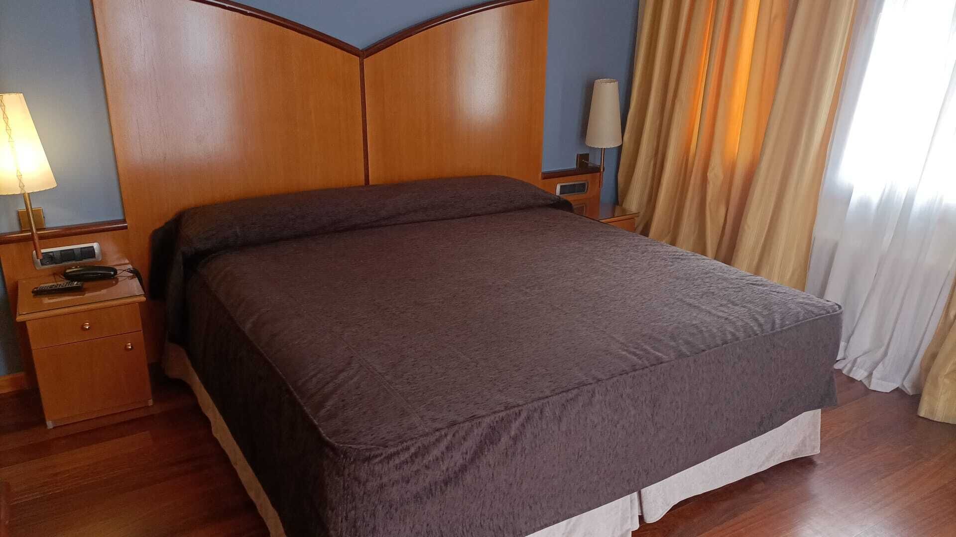 Een hotelkamer met een houten hoofdeinde, een bed met een bruine deken, een nachtkastje met een lamp en een raam met gordijnen.