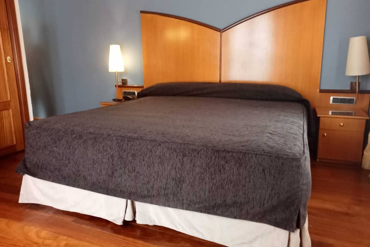 A neatly made bed with a grey bedspread and white skirted legs, a wooden headboard, and bedside tables with lamps.