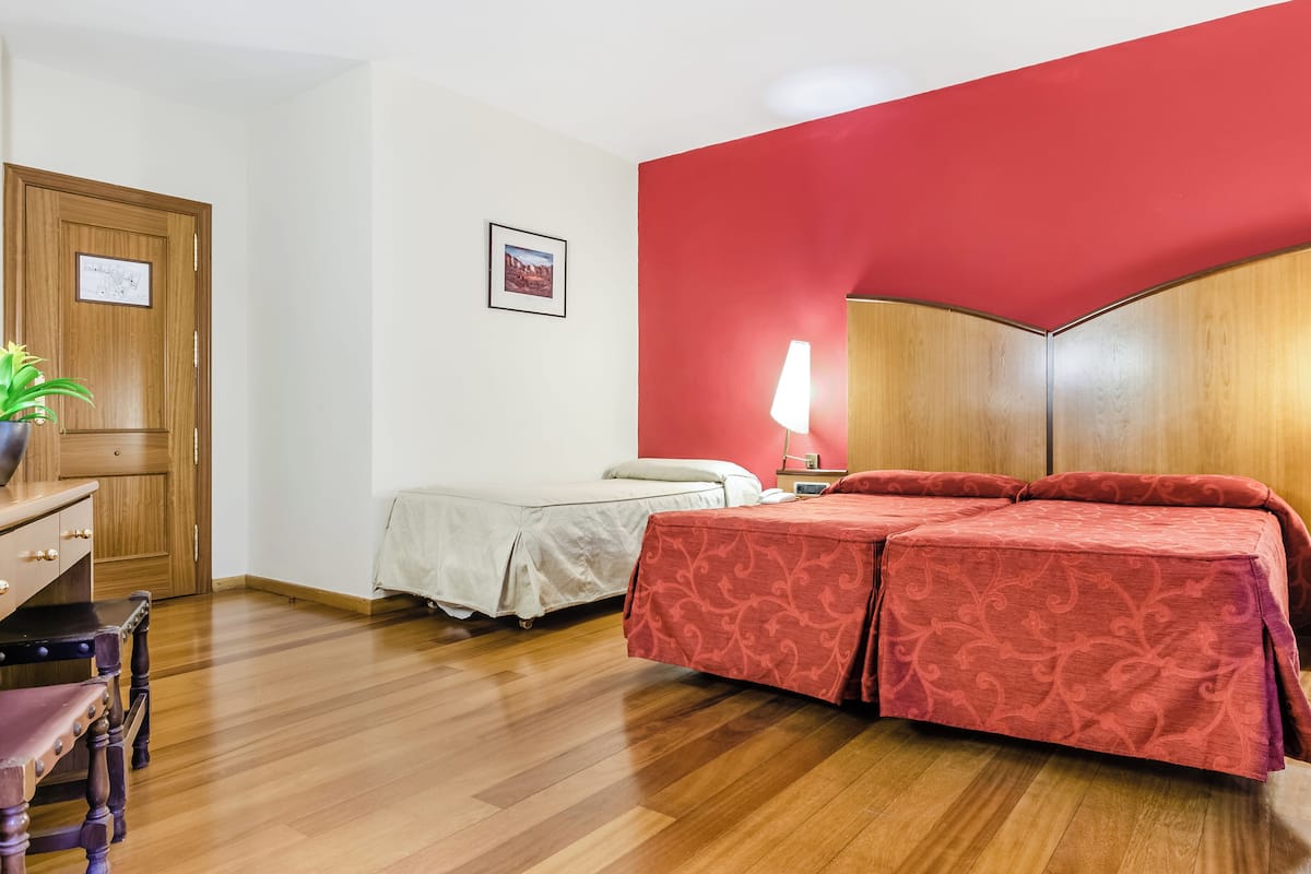 A room with a wooden desk, a bed with a red bedspread, a nightstand with a lamp, and a framed picture on the wall.