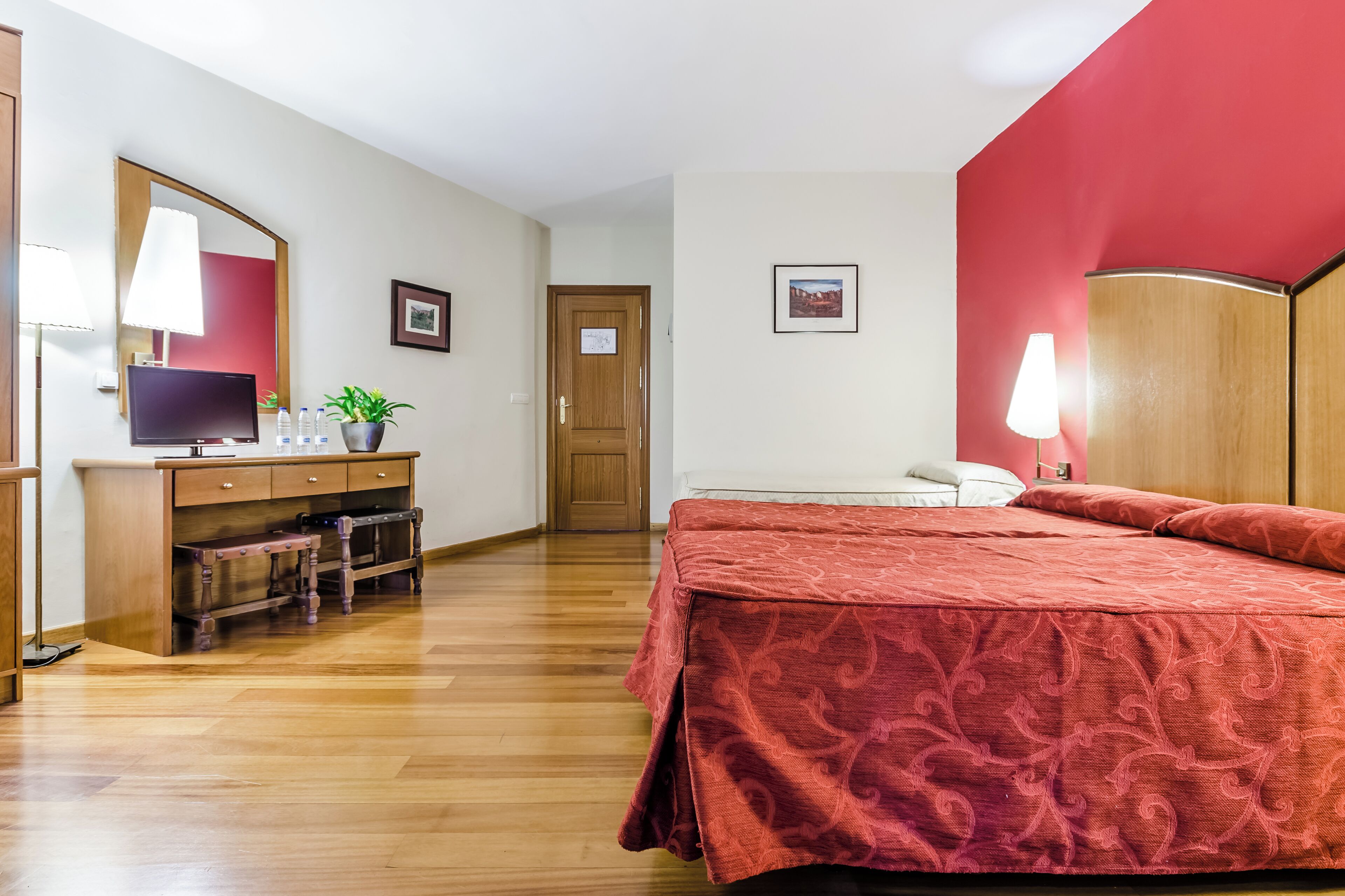 A hotel room with a wooden desk, a bed with a red bedspread, a mirror, and a door.