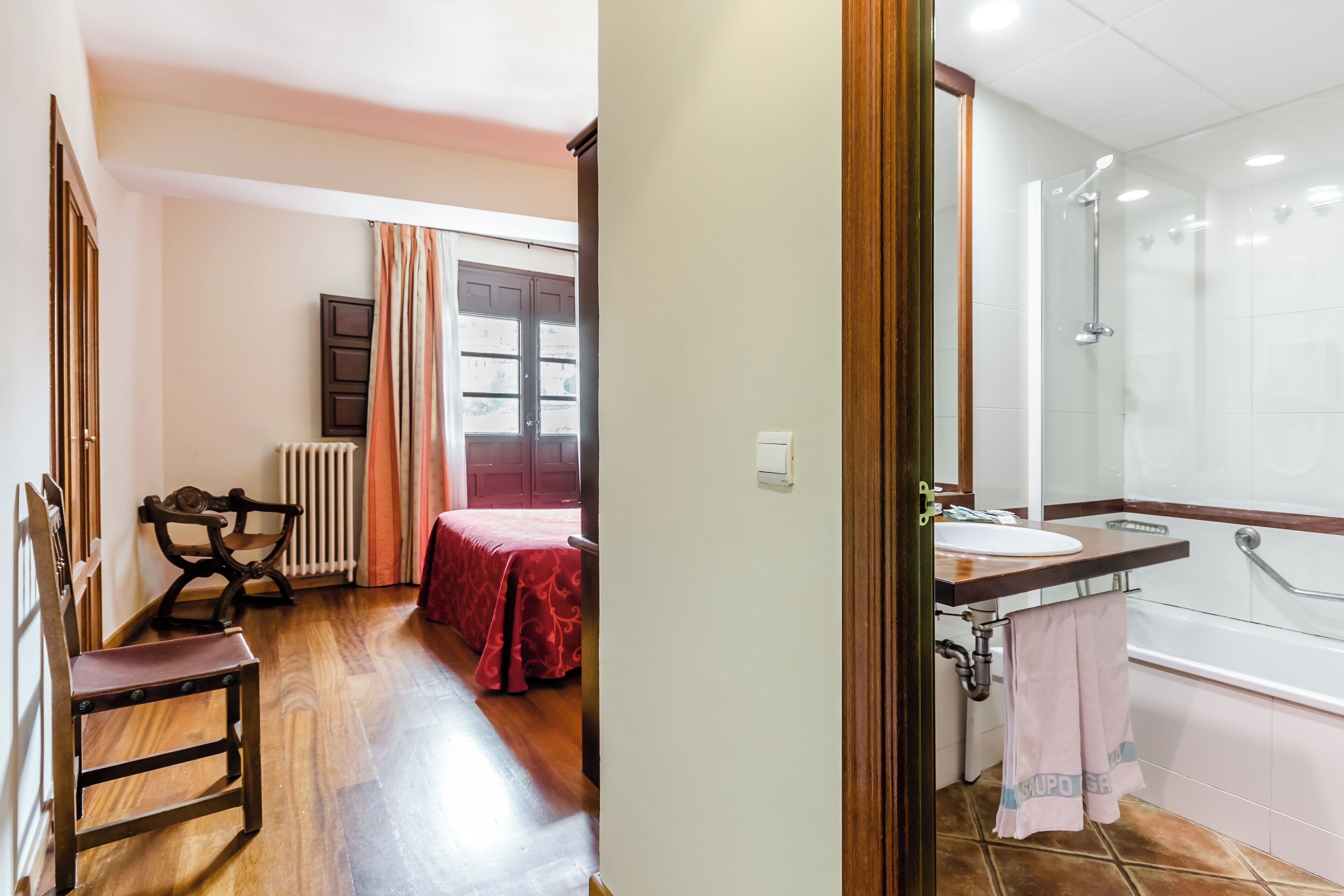 A room with a bed, a chair, a radiator, a window with curtains, and a bathroom with a sink and shower.