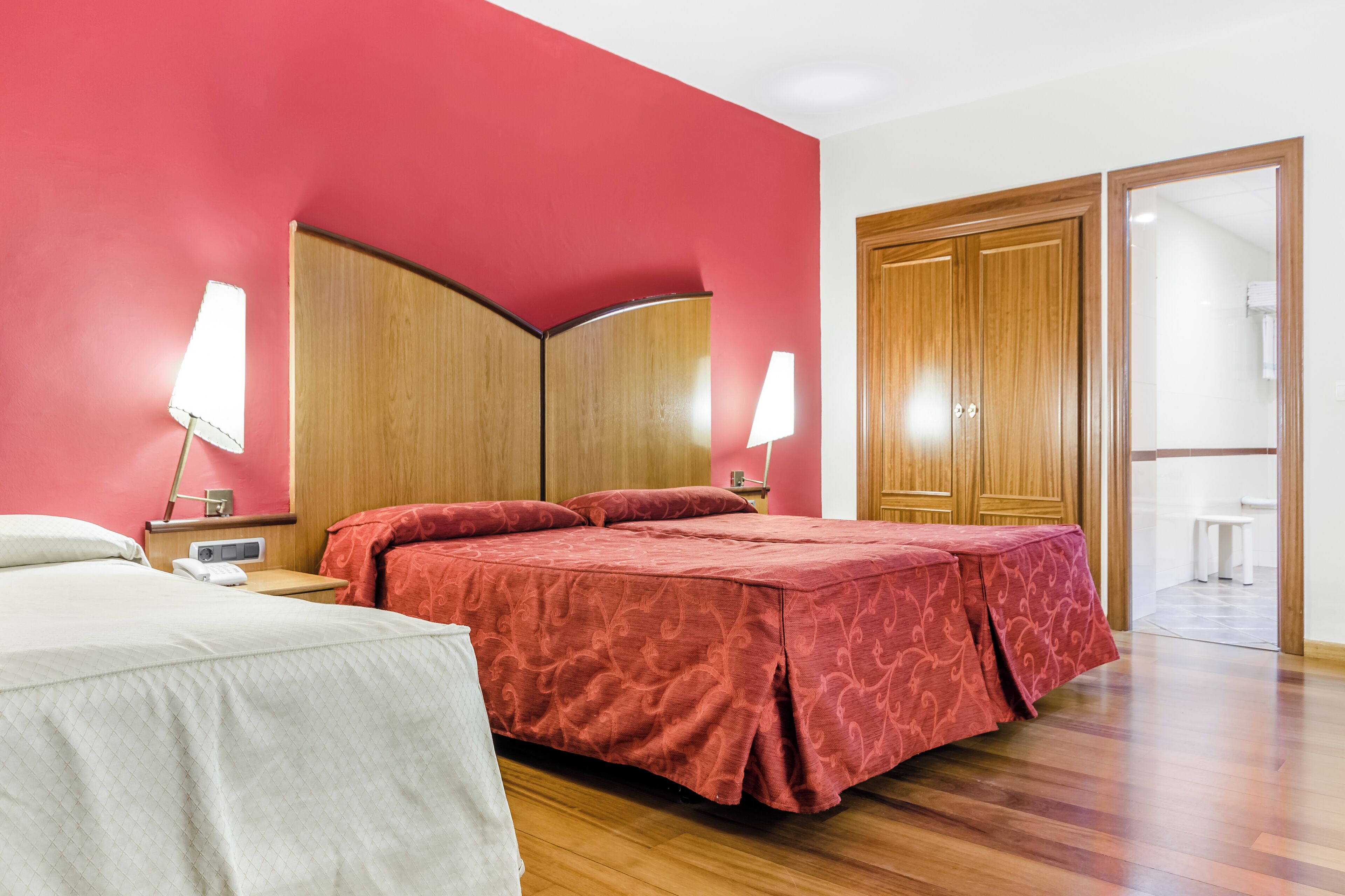 Habitación de hotel con cama, mesitas de noche, lámparas, un armario de madera y un baño visible a través de una puerta abierta.
