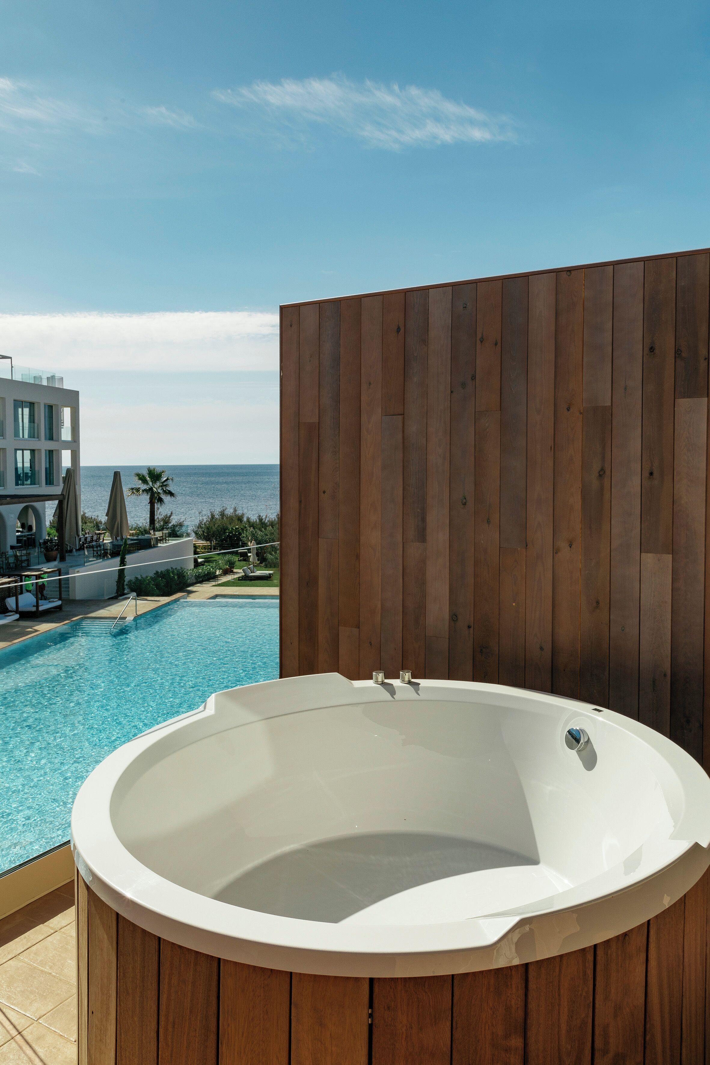 A hot tub with a wooden wall backdrop and a pool in the background.