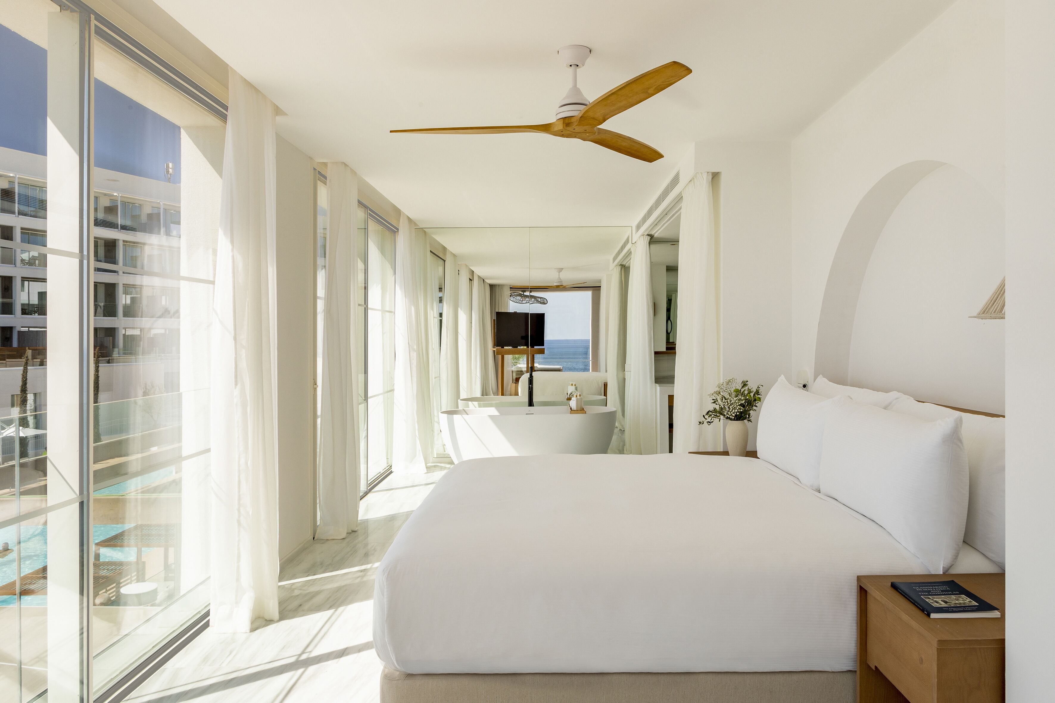A modern bedroom with a large bed, a ceiling fan, and a view of a pool and building through large windows.