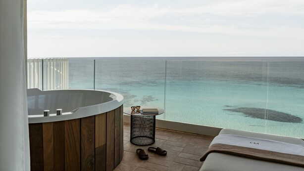 A balcony with a view of the ocean, a wooden floor, a small round table with a book and glasses, and a pair of slippers.
