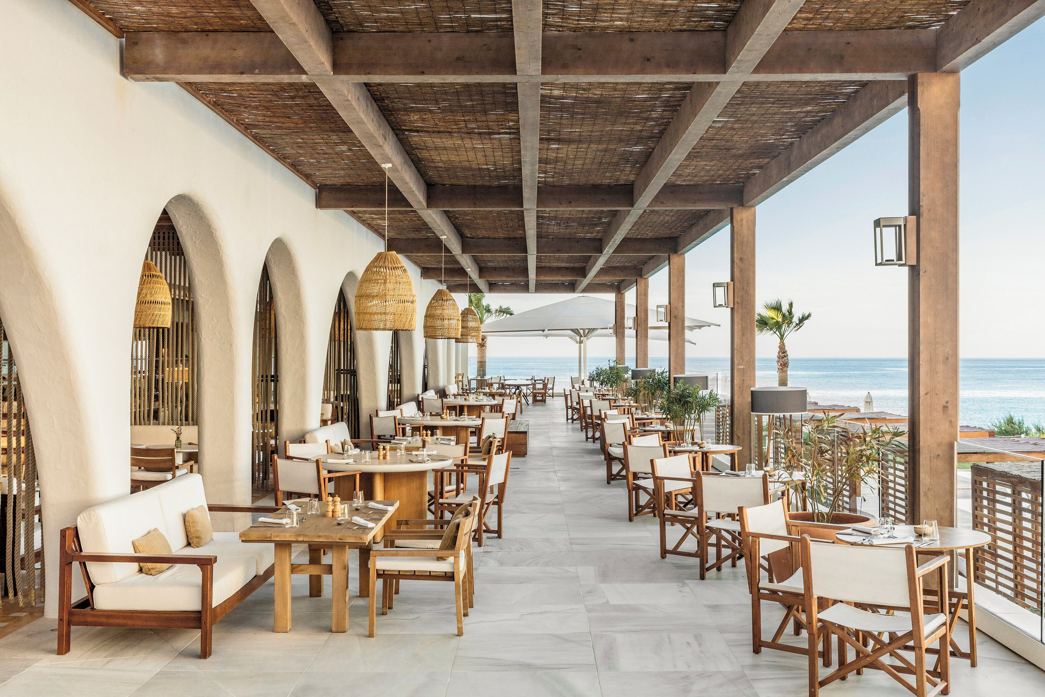 A spacious outdoor dining area with wooden tables and chairs, a white sofa, and a view of the ocean.
