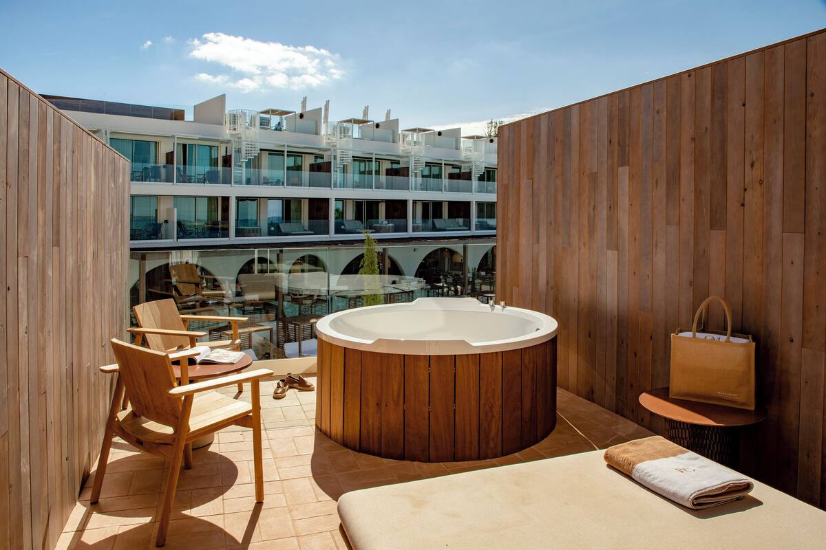 Un espace de détente extérieur moderne, doté d’une terrasse en bois, d’un jacuzzi, de chaises longues et offrant une vue sur un immeuble à plusieurs étages avec des balcons.