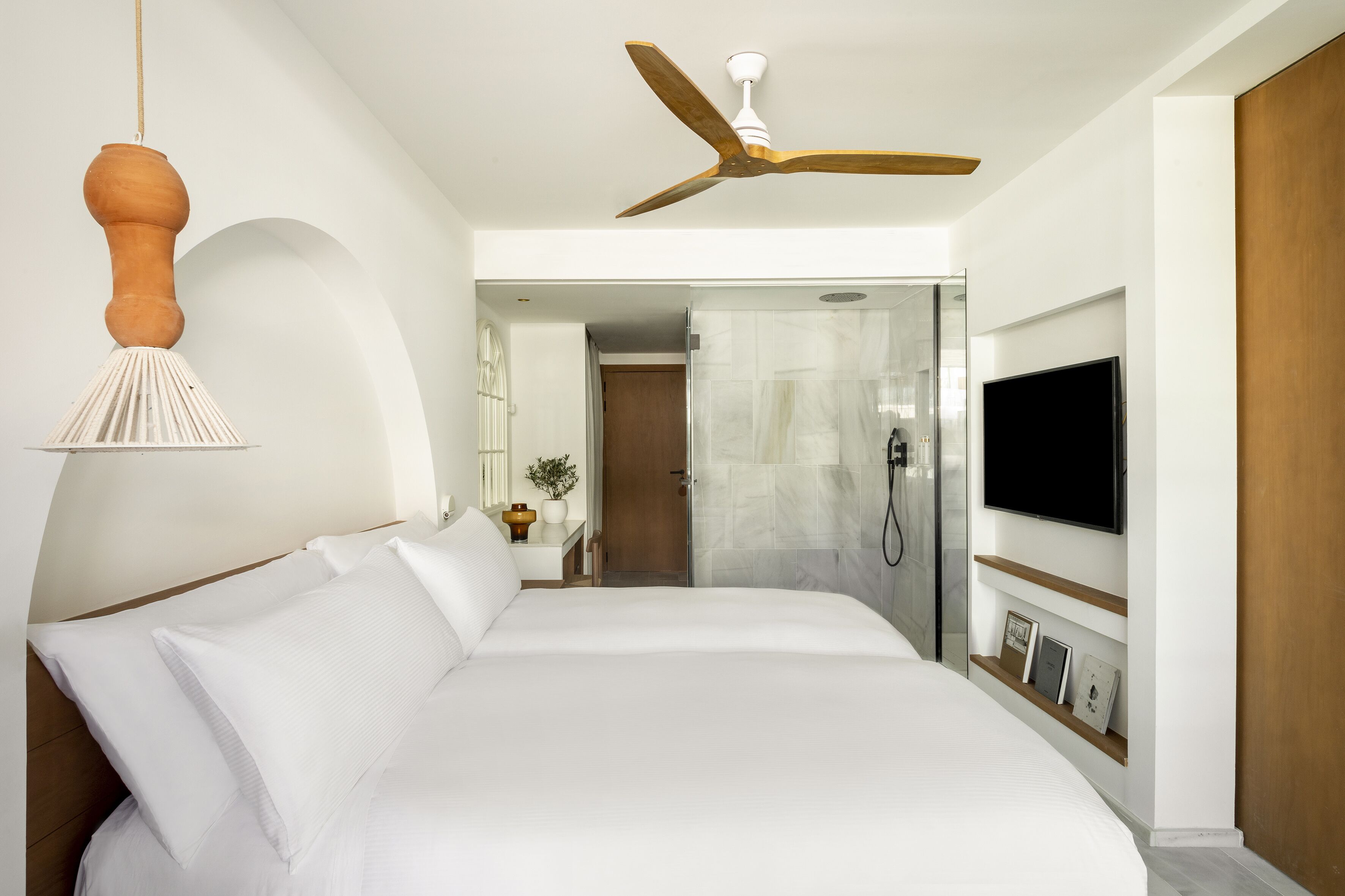 Ein Hotelzimmer mit einem großen Bett, einem Deckenventilator, einem an der Wand befestigten Fernseher und einem Duschbereich.