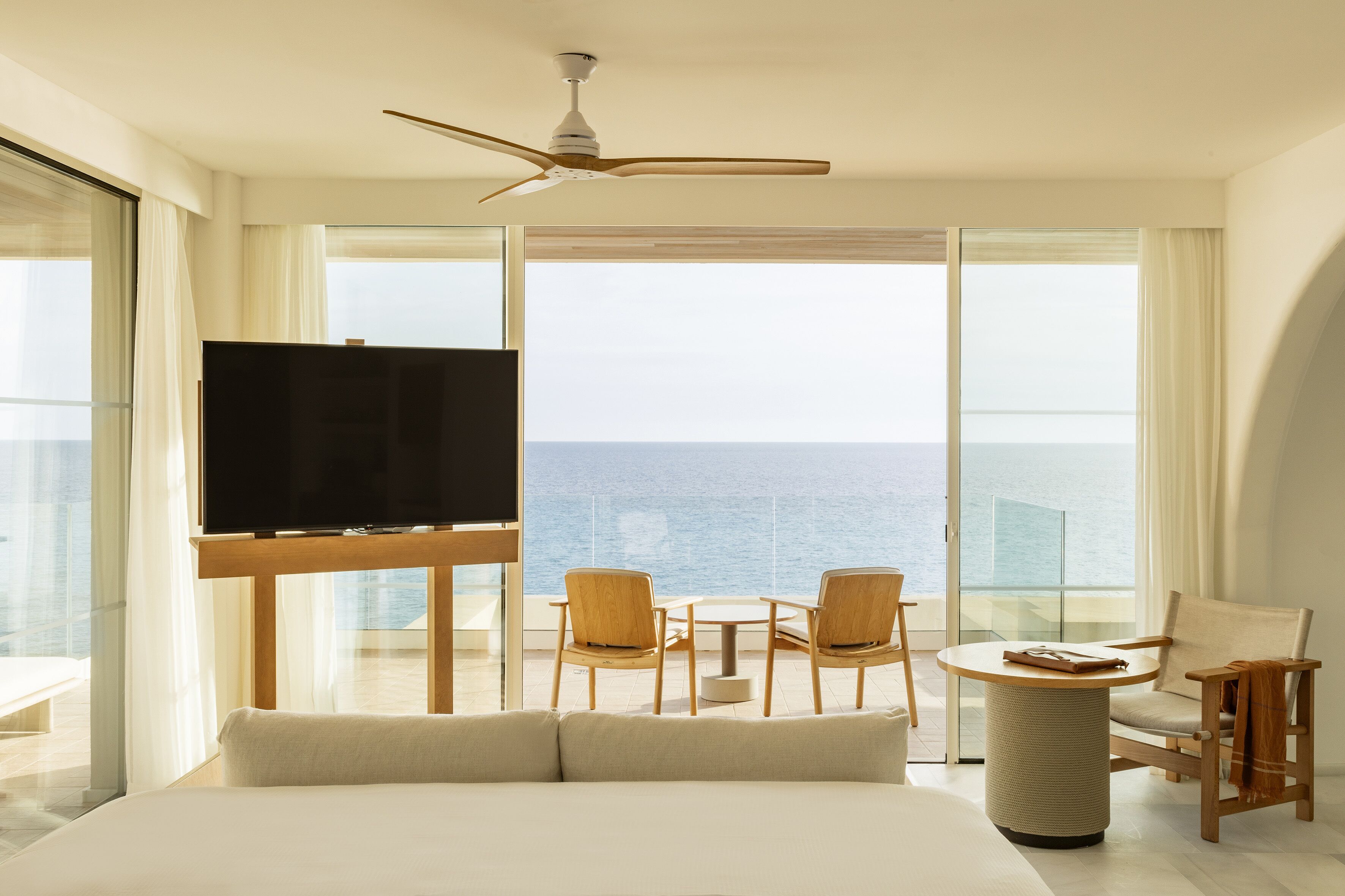 Habitació d'hotel amb un llit gran, un televisor sobre un suport de fusta, un balcó amb vista al mar i una taula petita amb cadires.