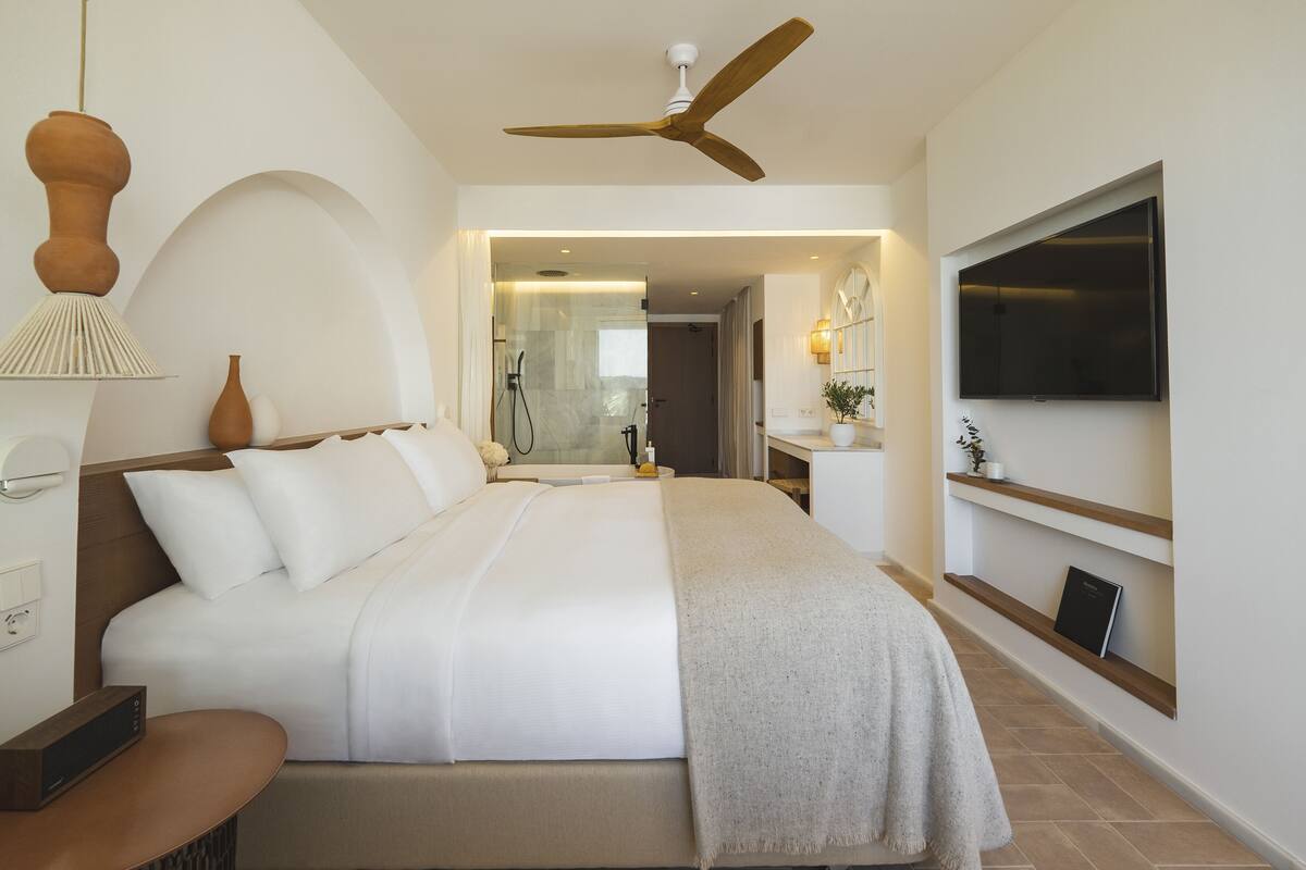 Une chambre d’hôtel moderne avec un grand lit, un ventilateur de plafond, un téléviseur fixé au mur et une salle de bain visible à travers une porte ouverte.
