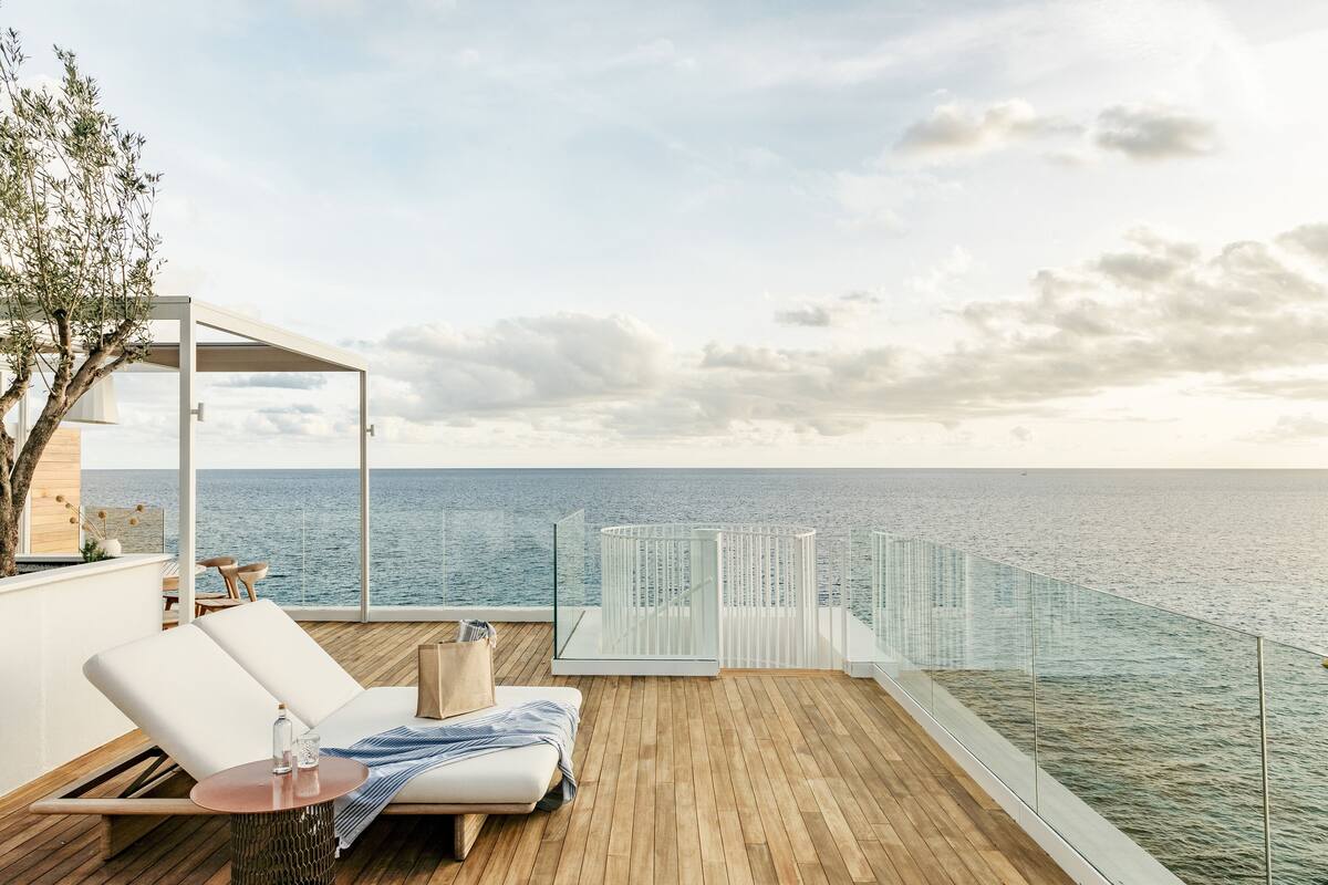 Une terrasse en bois dotée d’une balustrade en verre donnant sur la mer, une chaise longue blanche et une petite table sur laquelle se trouvent un bocal en verre et un livre.