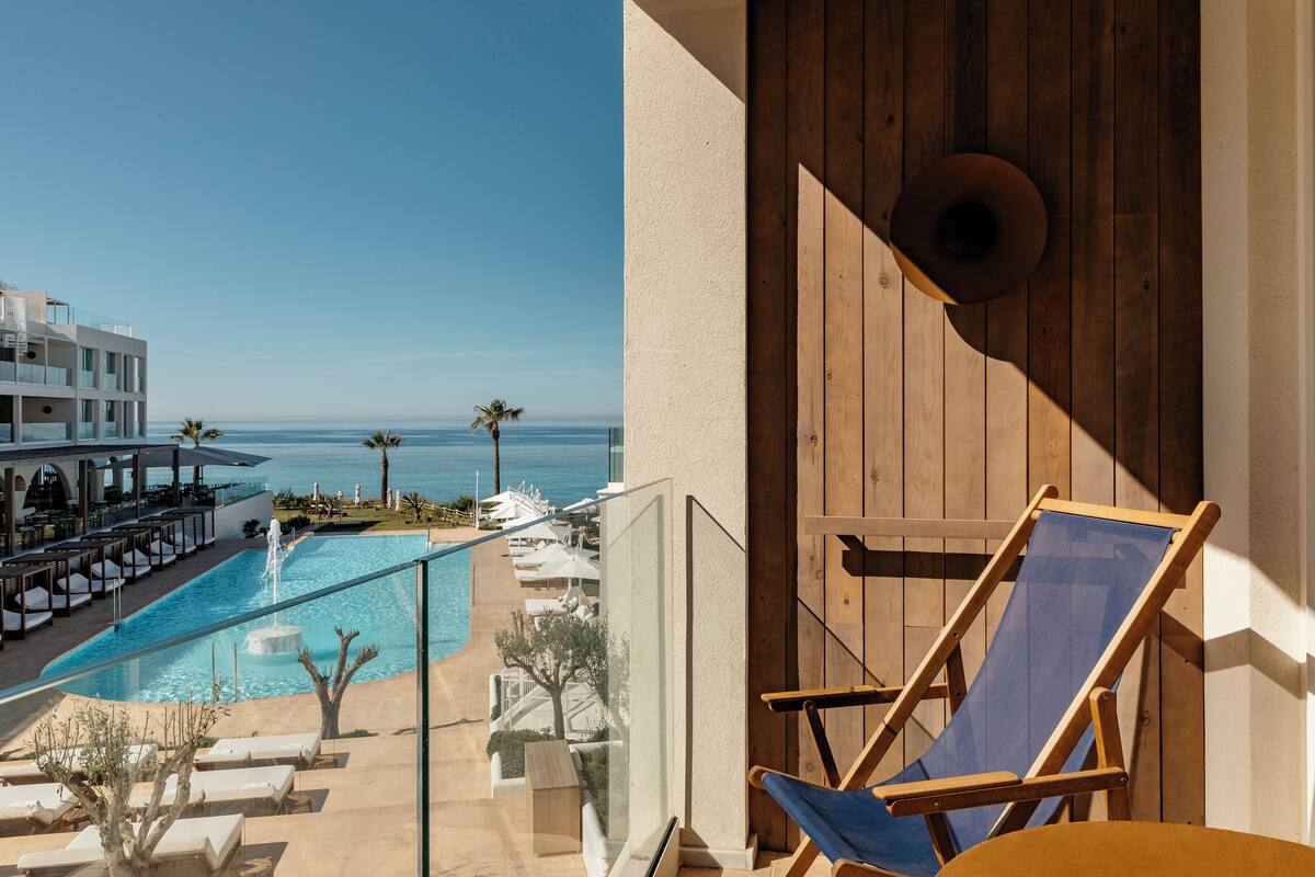 Un balcon avec vue sur une piscine et la mer, des chaises longues en bois et un espace détente avec des parasols.