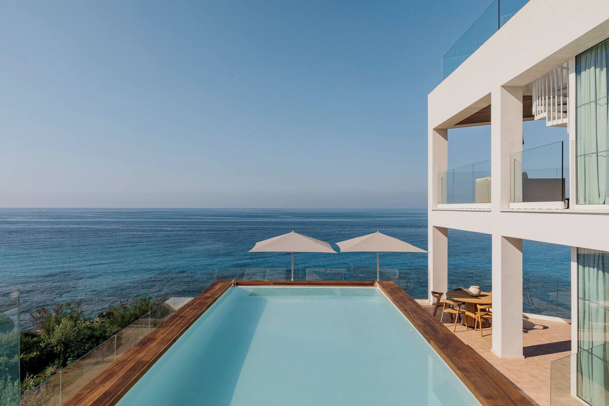 Une piscine moderne offrant une vue dégagée sur l’océan, une terrasse en bois et un espace balcon vitré aménagé avec du mobilier d’extérieur.