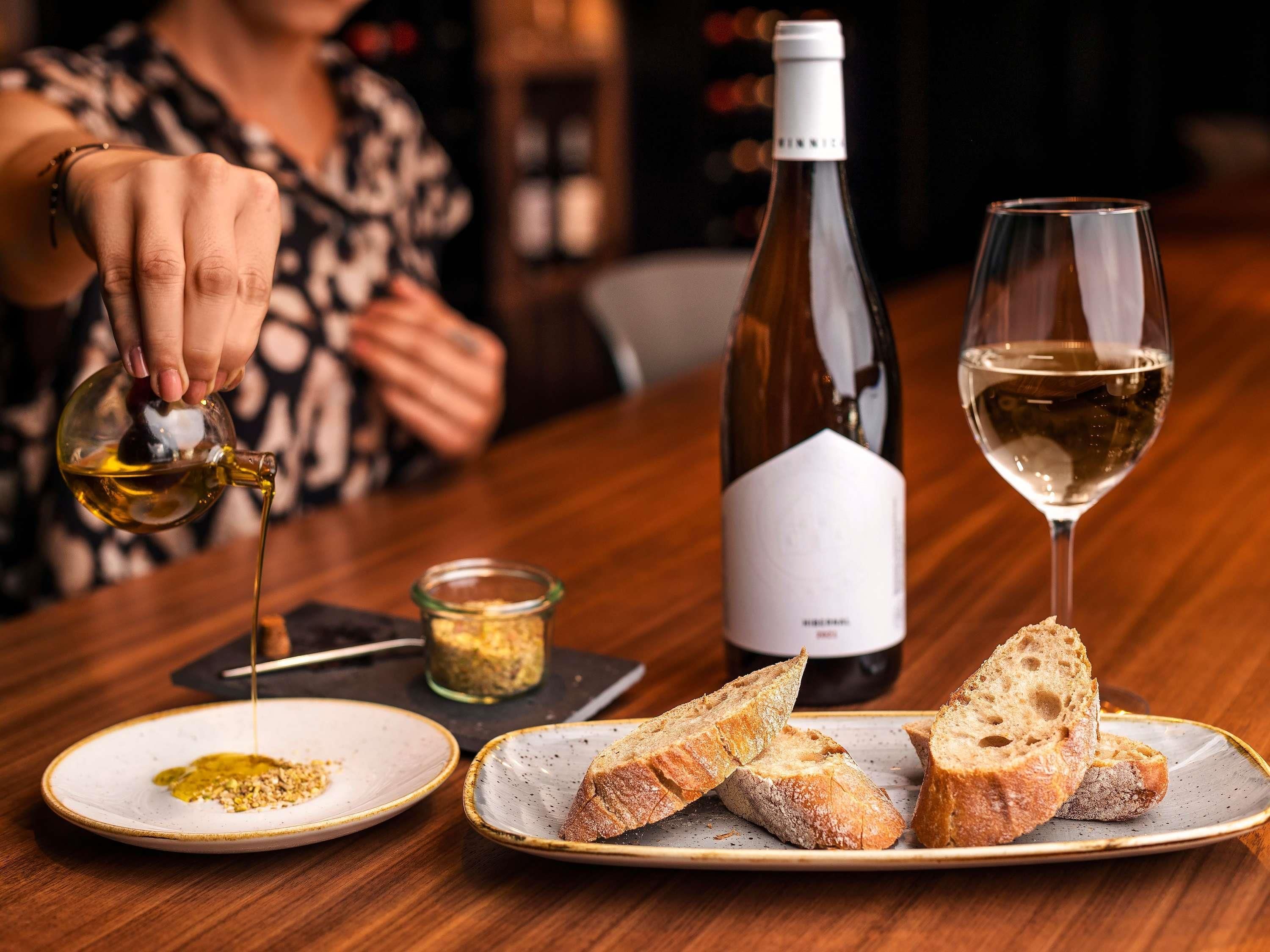 A person pouring olive oil onto bread, a bottle of wine, and a glass of wine on a table.