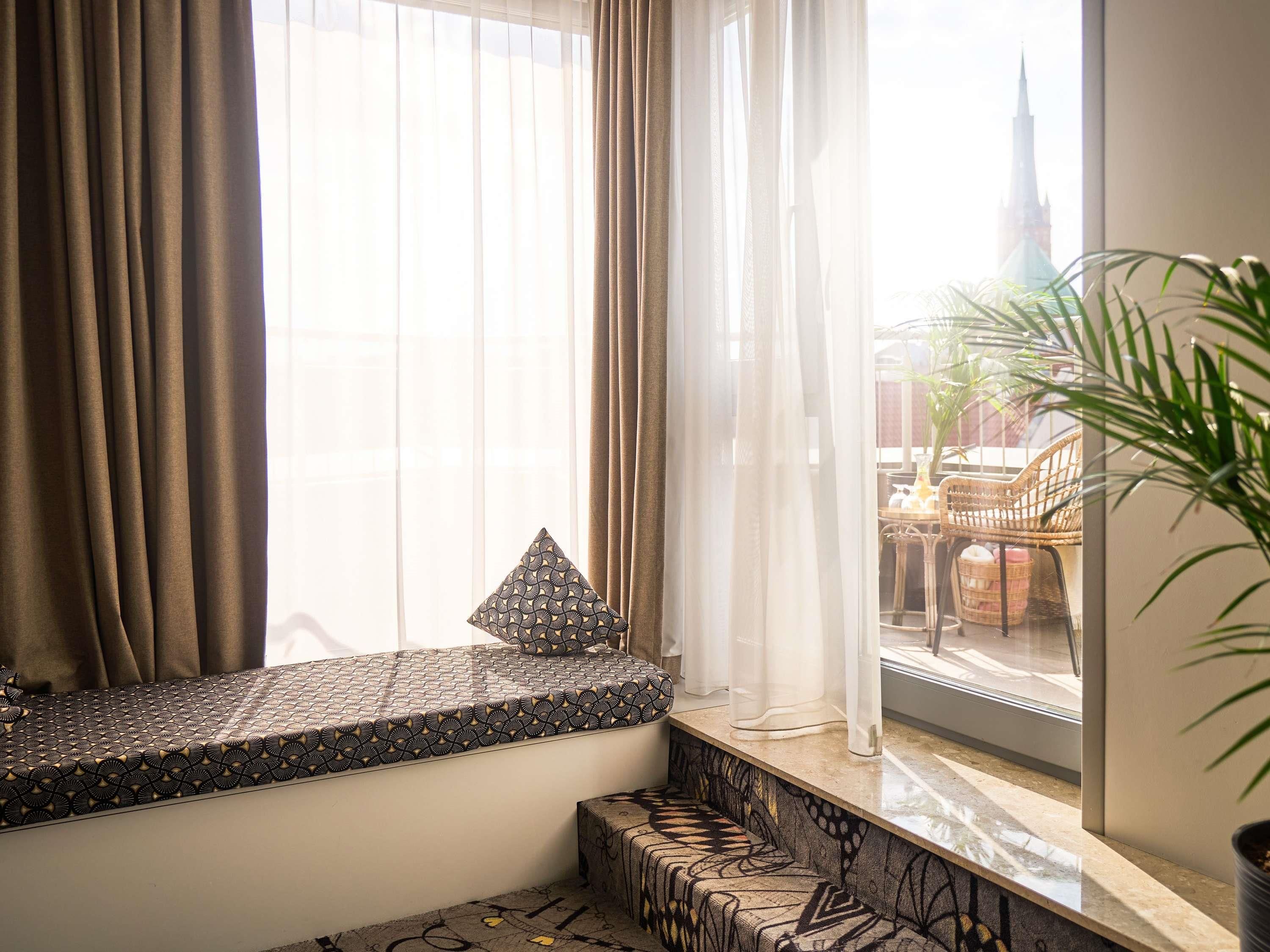 Habitación con ventana que ofrece vistas a un paisaje urbano, con un banco acolchado y un cojín estampado.