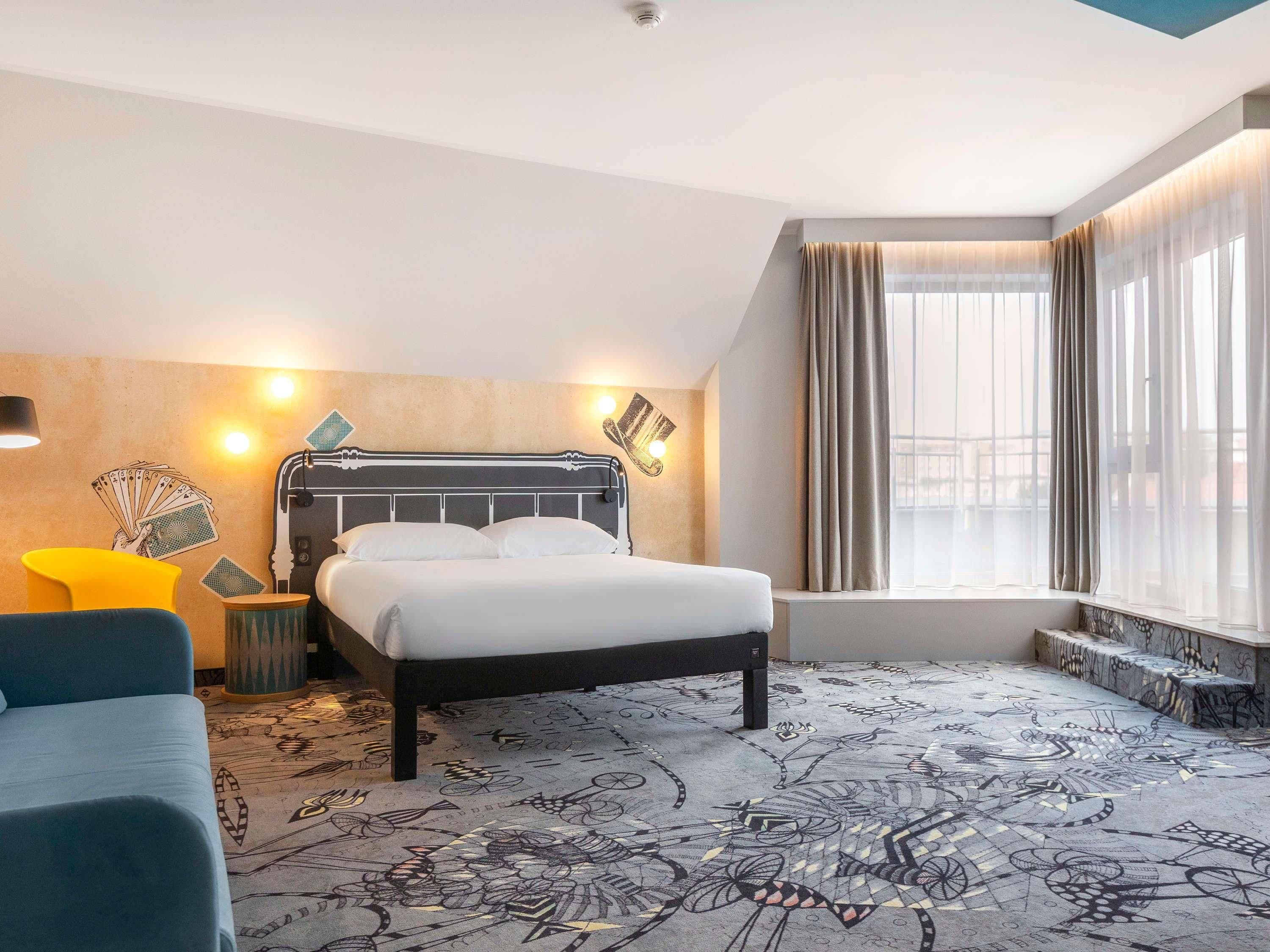 Een hotelkamer met een bed, een stoel, een nachtkastje en een raam met gordijnen.