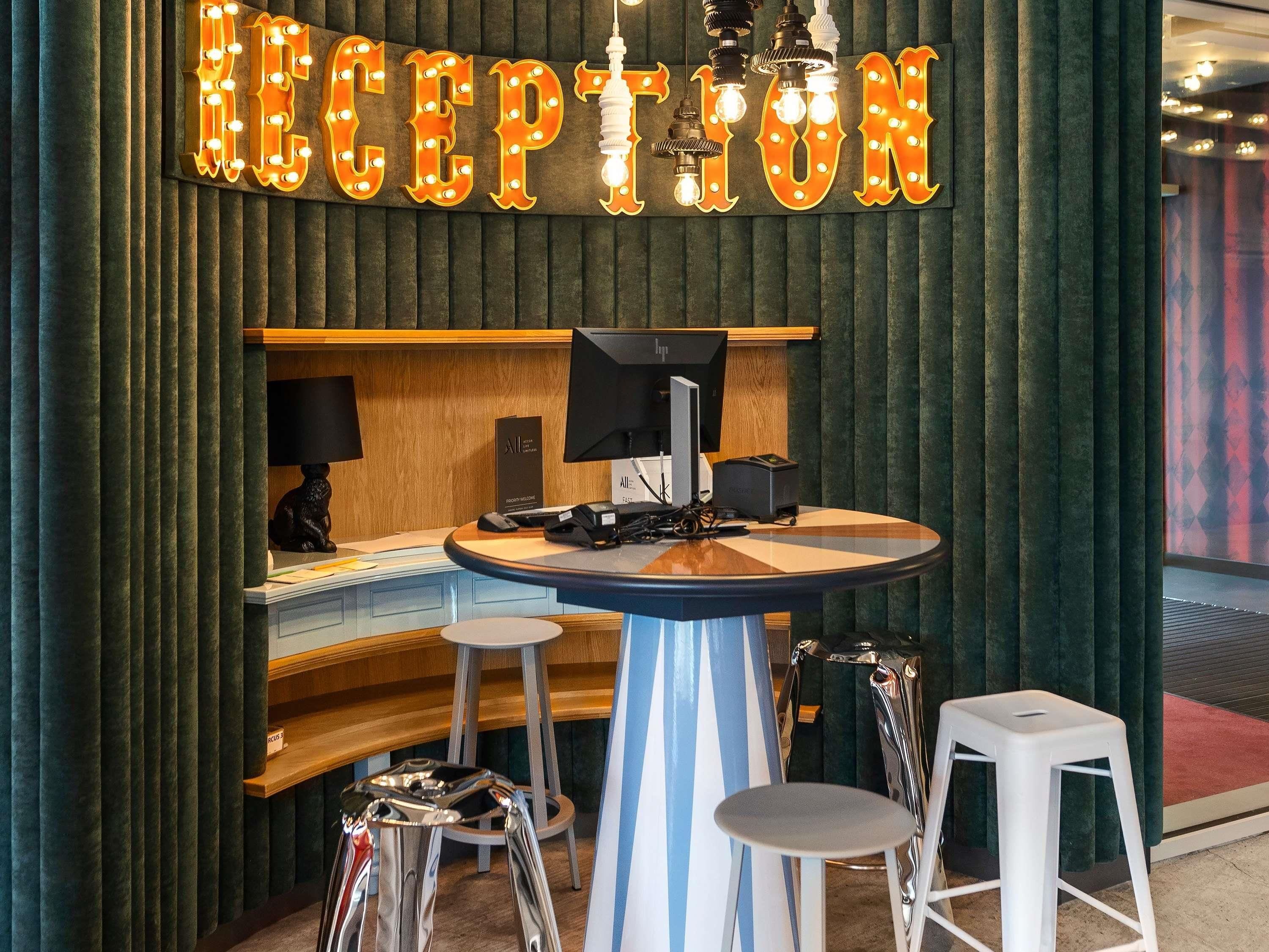 Een receptiegedeelte met een bord waarop 'RECEPTION' staat, een bureau met een computer, telefoon en een klein zitgedeelte met krukjes.
