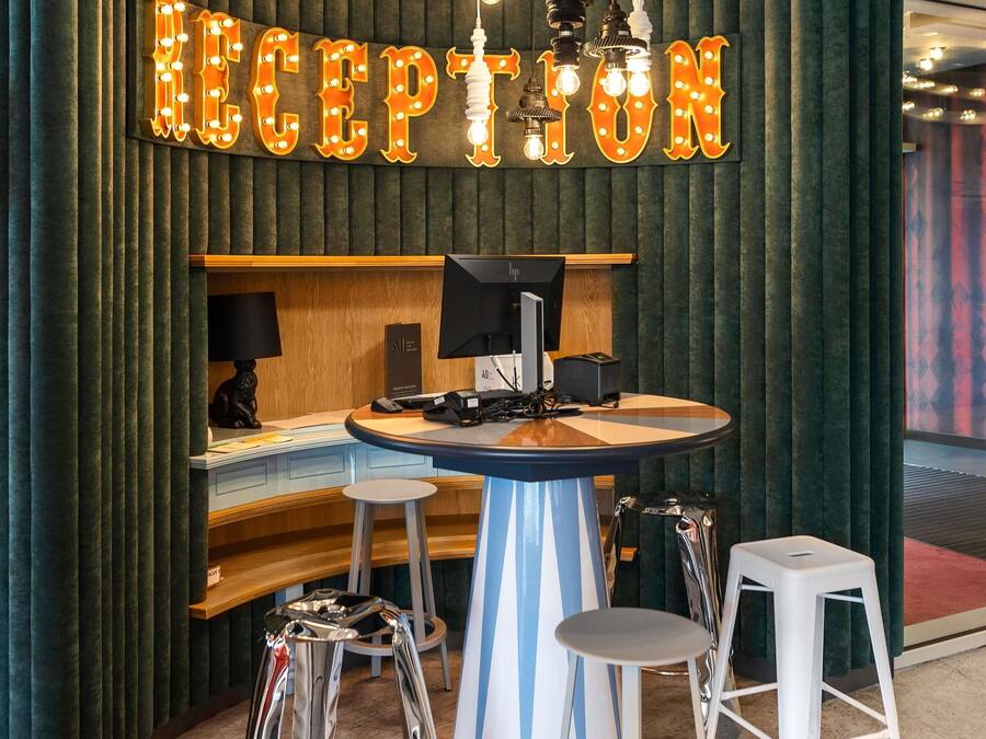 Een receptiegedeelte met een bord waarop 'RECEPTION' staat, een bureau met een computer, telefoon en een klein zitgedeelte met krukjes.