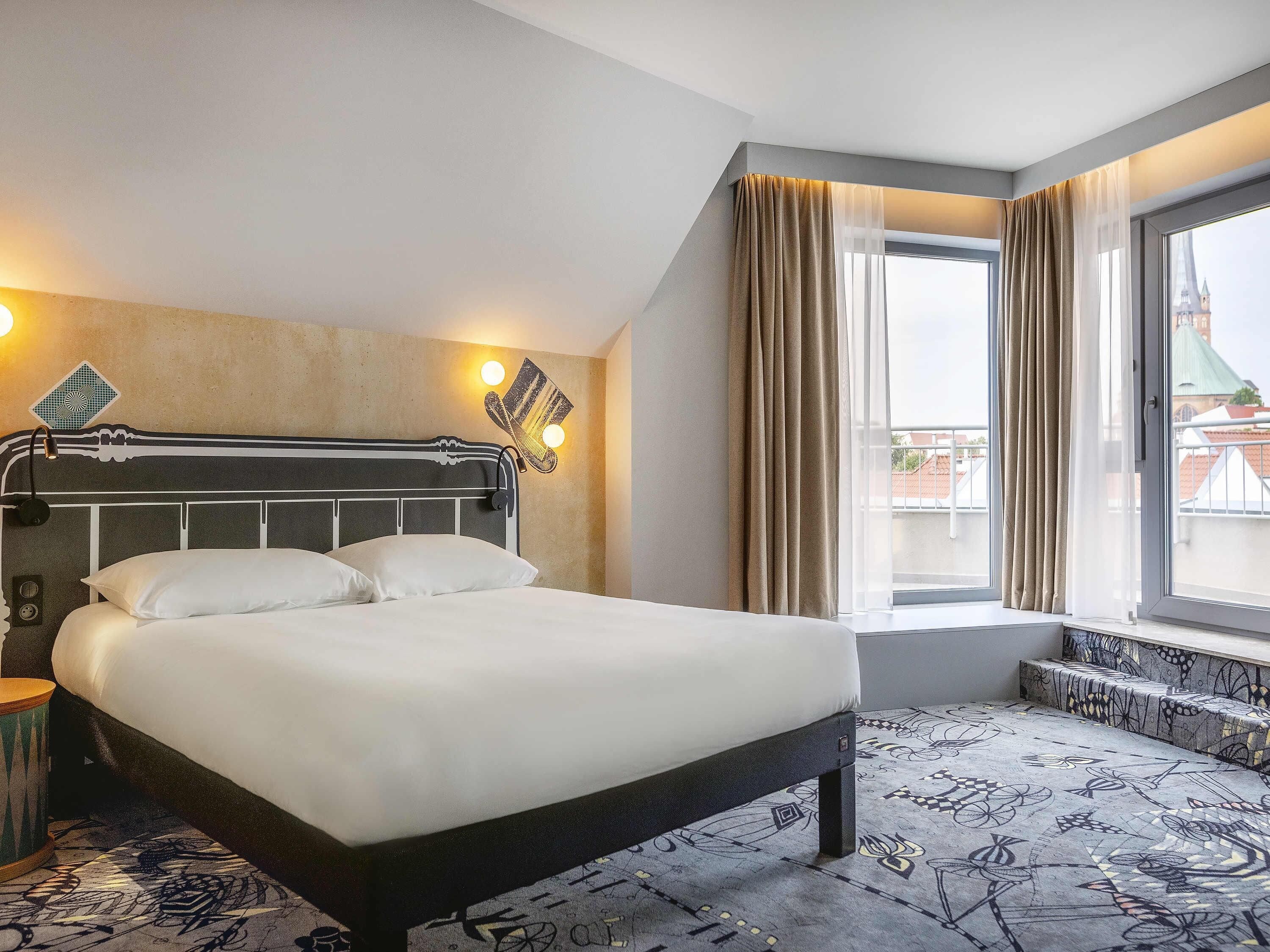 Una camera d'albergo con un letto grande, una testiera con luci a parete, una finestra con tende e un tappeto decorato.