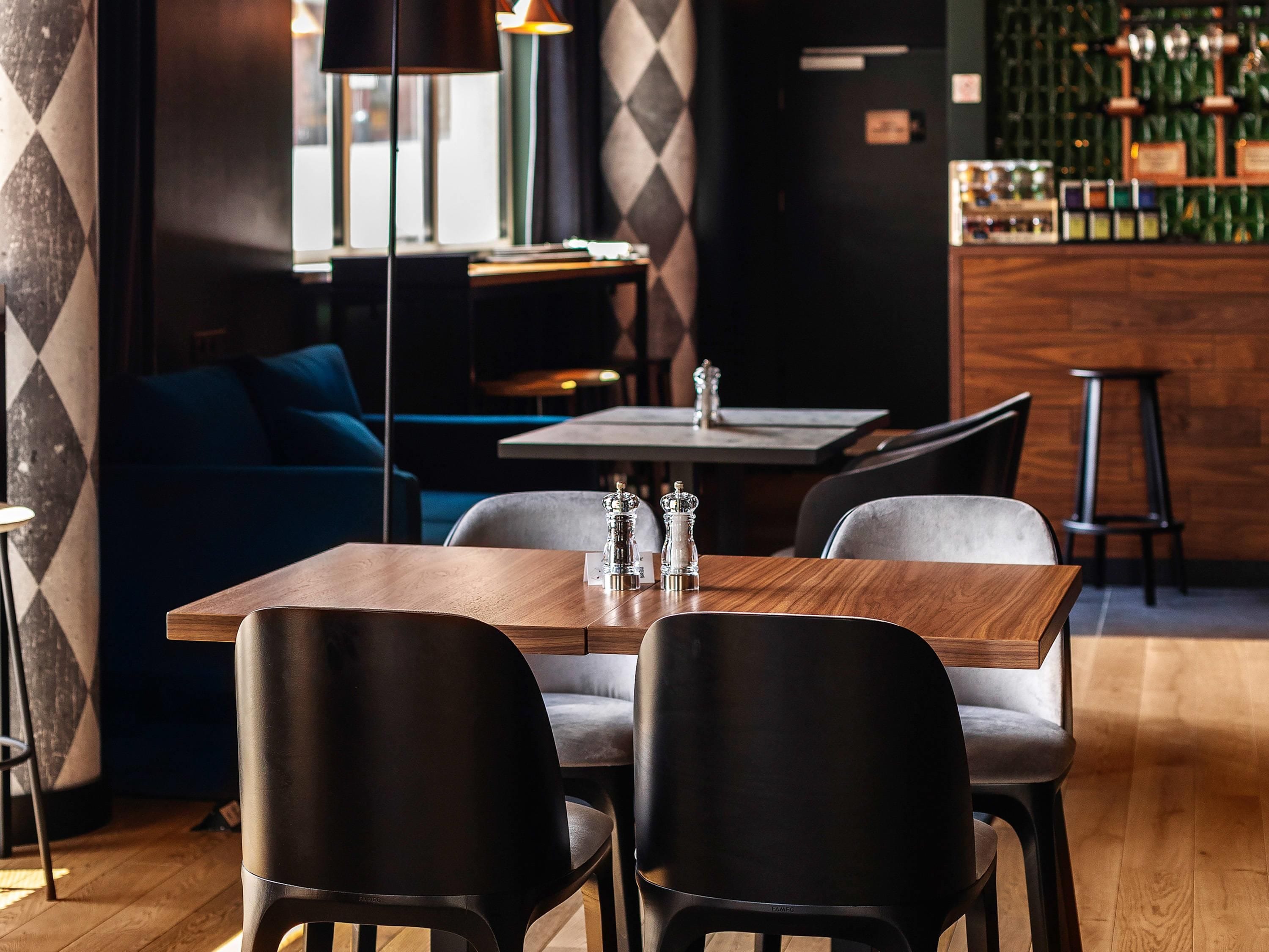 A modern restaurant interior with a wooden table, black chairs, a blue sofa, and a bar area with bottles.