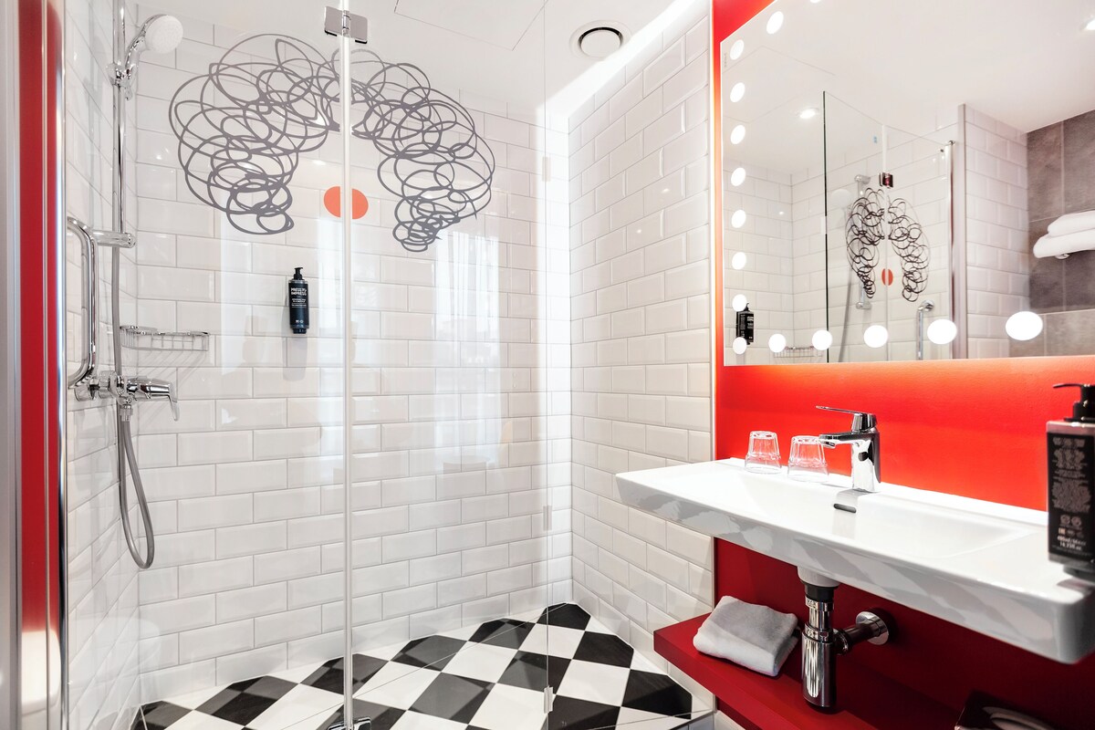 A modern bathroom with a glass-enclosed shower, a white sink with a red accent wall, and a checkered floor.