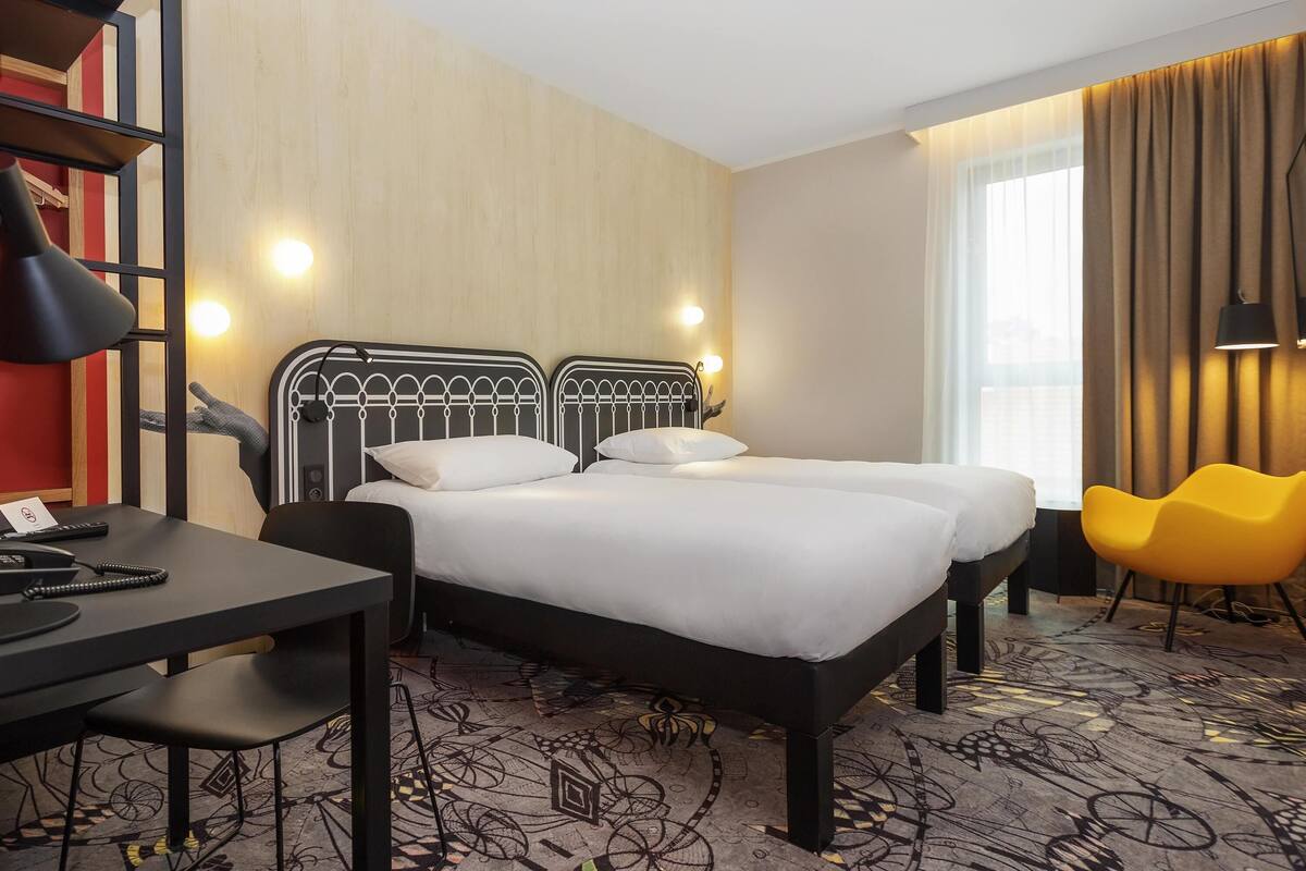 A hotel room with two beds, a desk, a chair, and a large window with curtains.