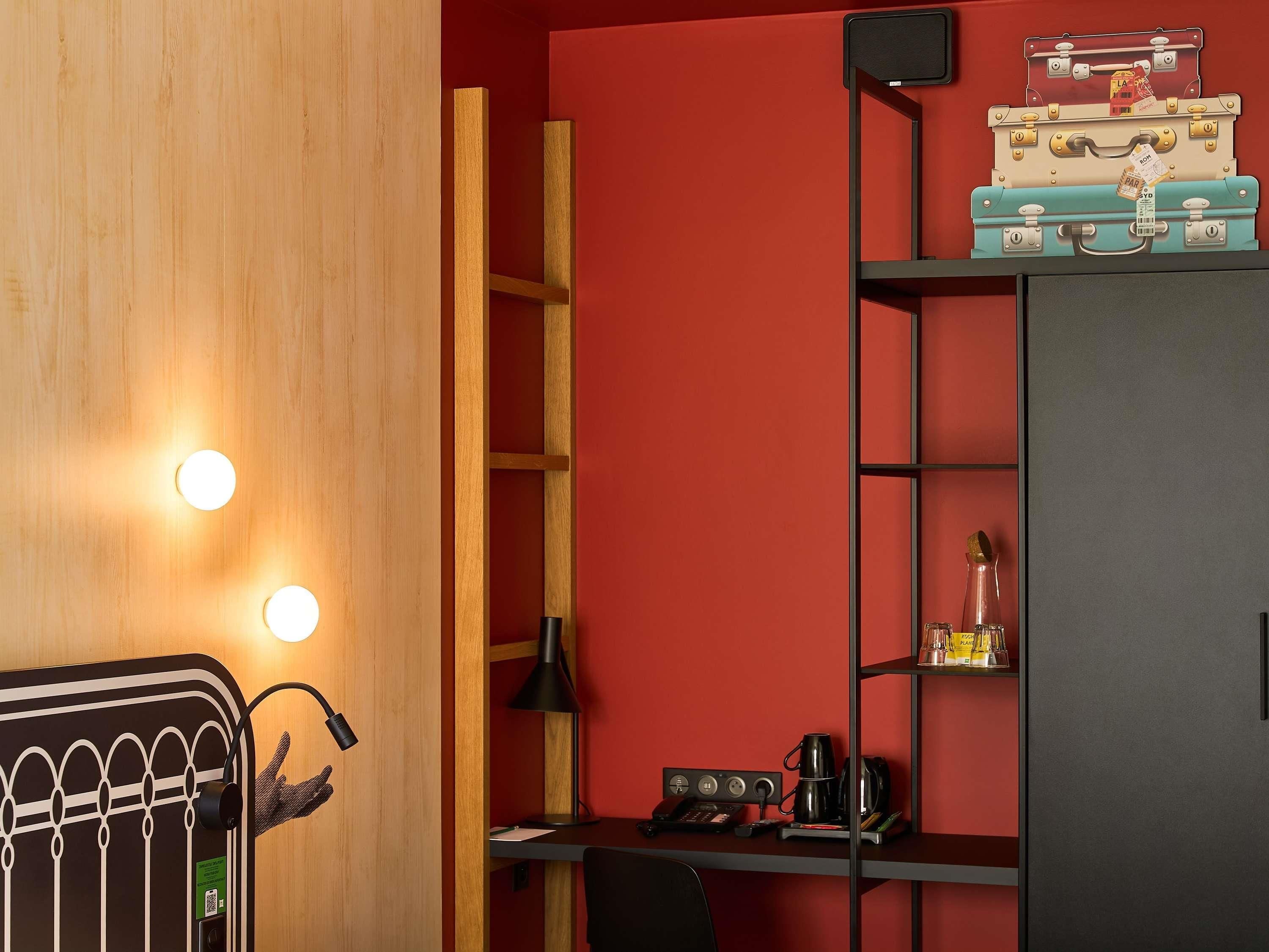 A room with a wooden door, a desk with a lamp, a shelf with suitcases, and a red wall.