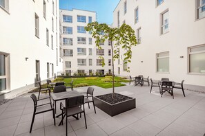 Un patio con mobiliario exterior, un pequeño árbol y un edificio al fondo.