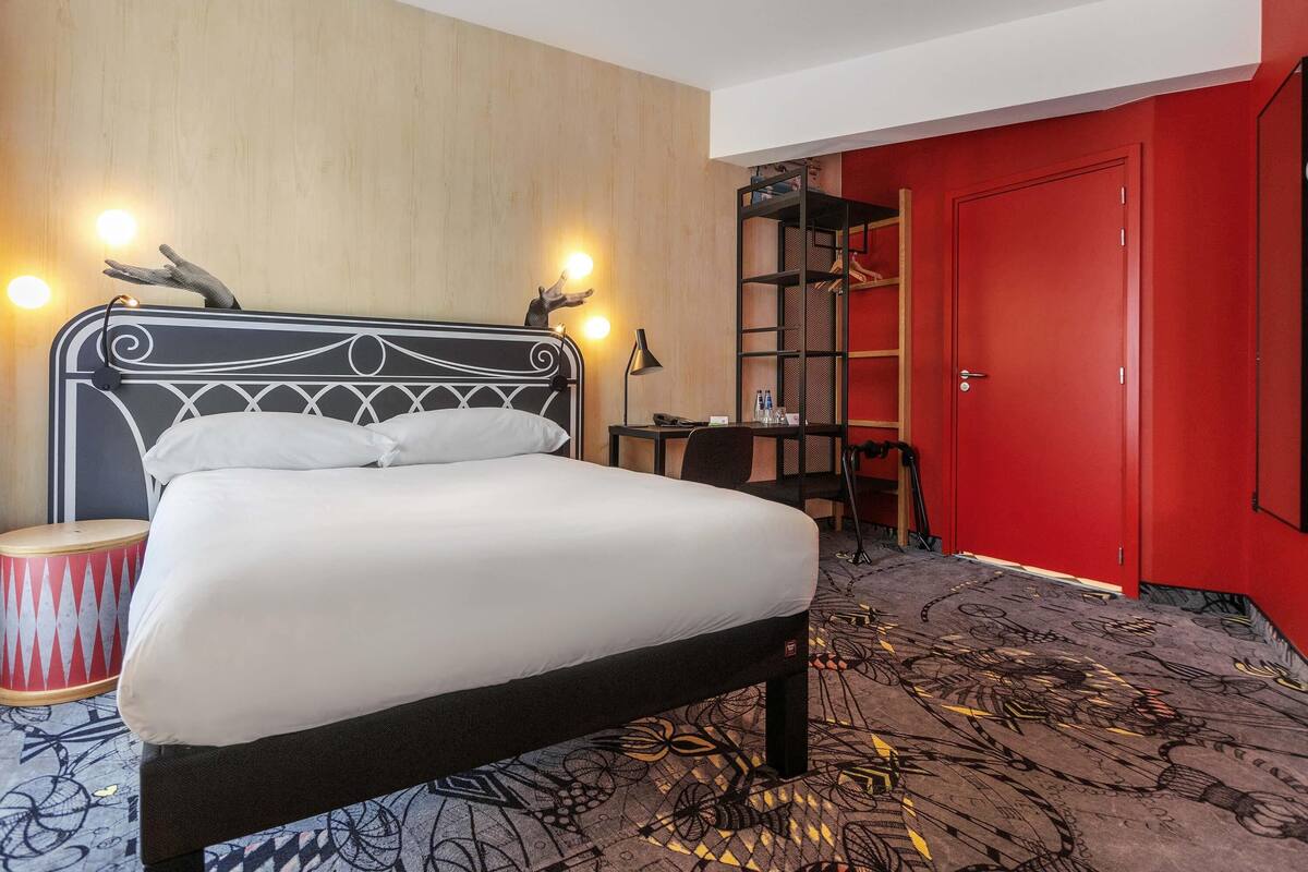 A hotel room with a bed, a desk, a chair, a wardrobe, and a red door.