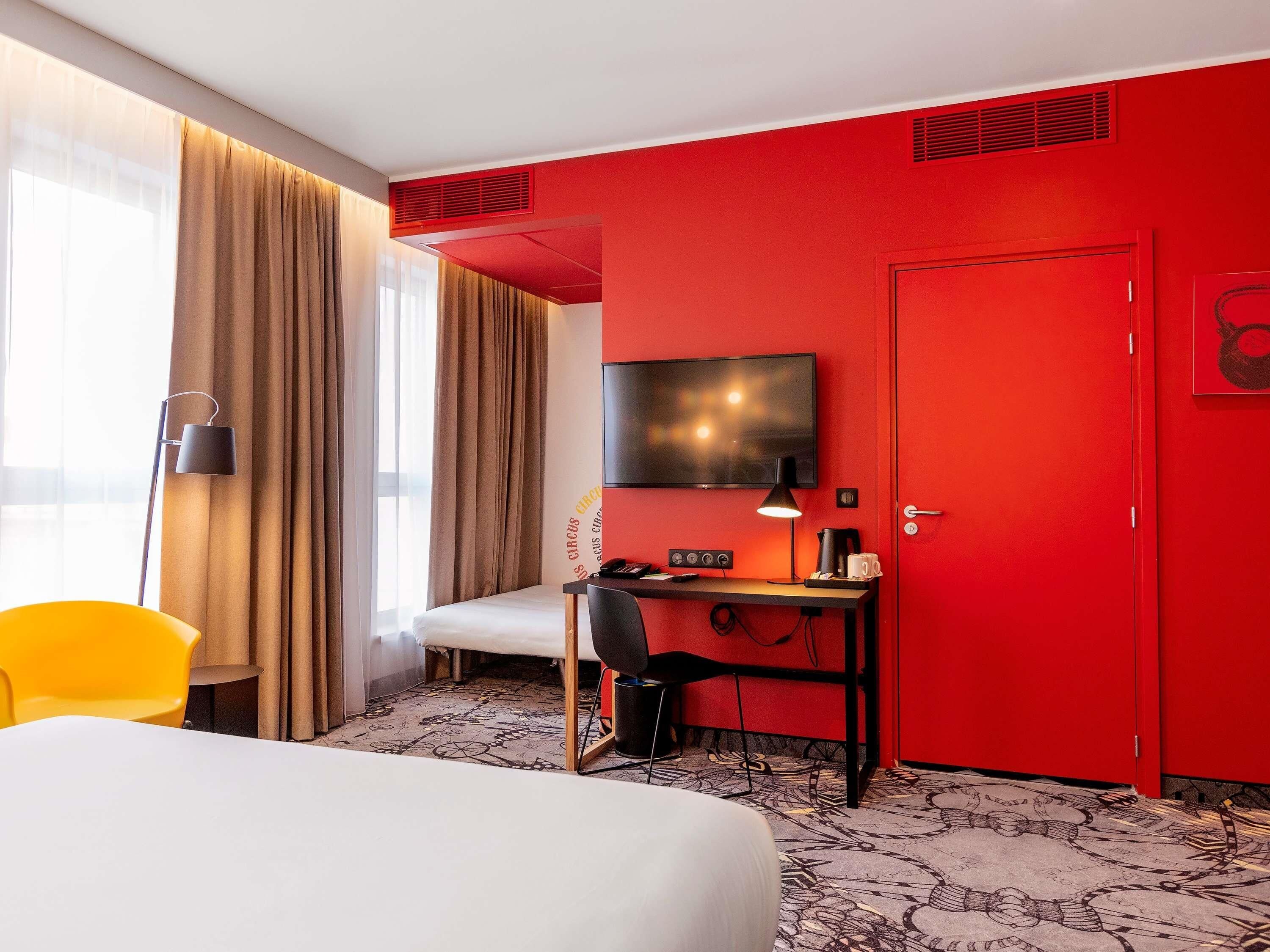 A hotel room with a bed, a desk with a chair, a television, and a red door.