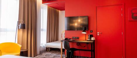 Quarto de hotel com cama, escrivaninha, cadeira, televisão e porta vermelha.