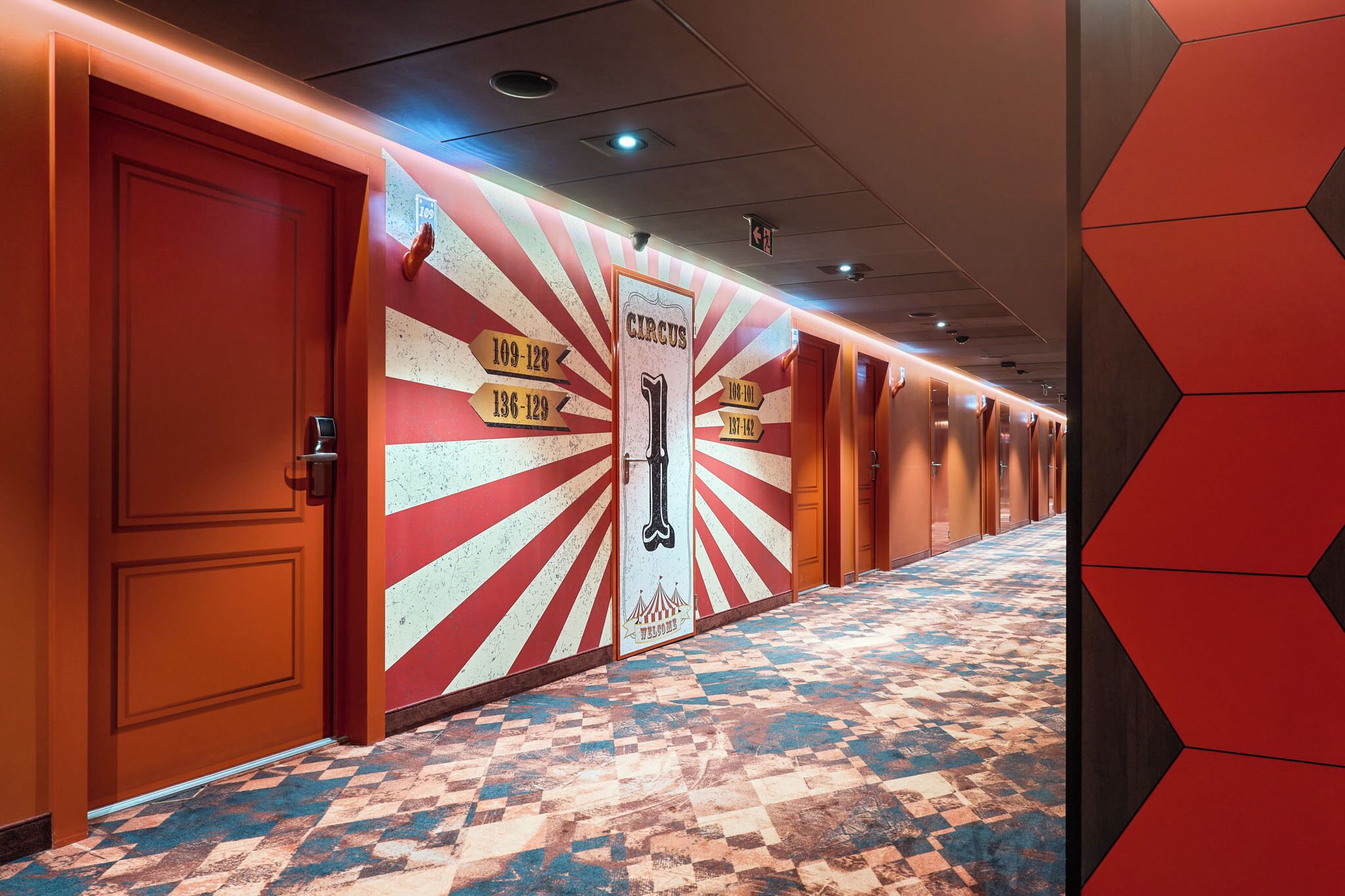 Un pasillo de hotel con una puerta roja, una pared con indicaciones y un suelo estampado.