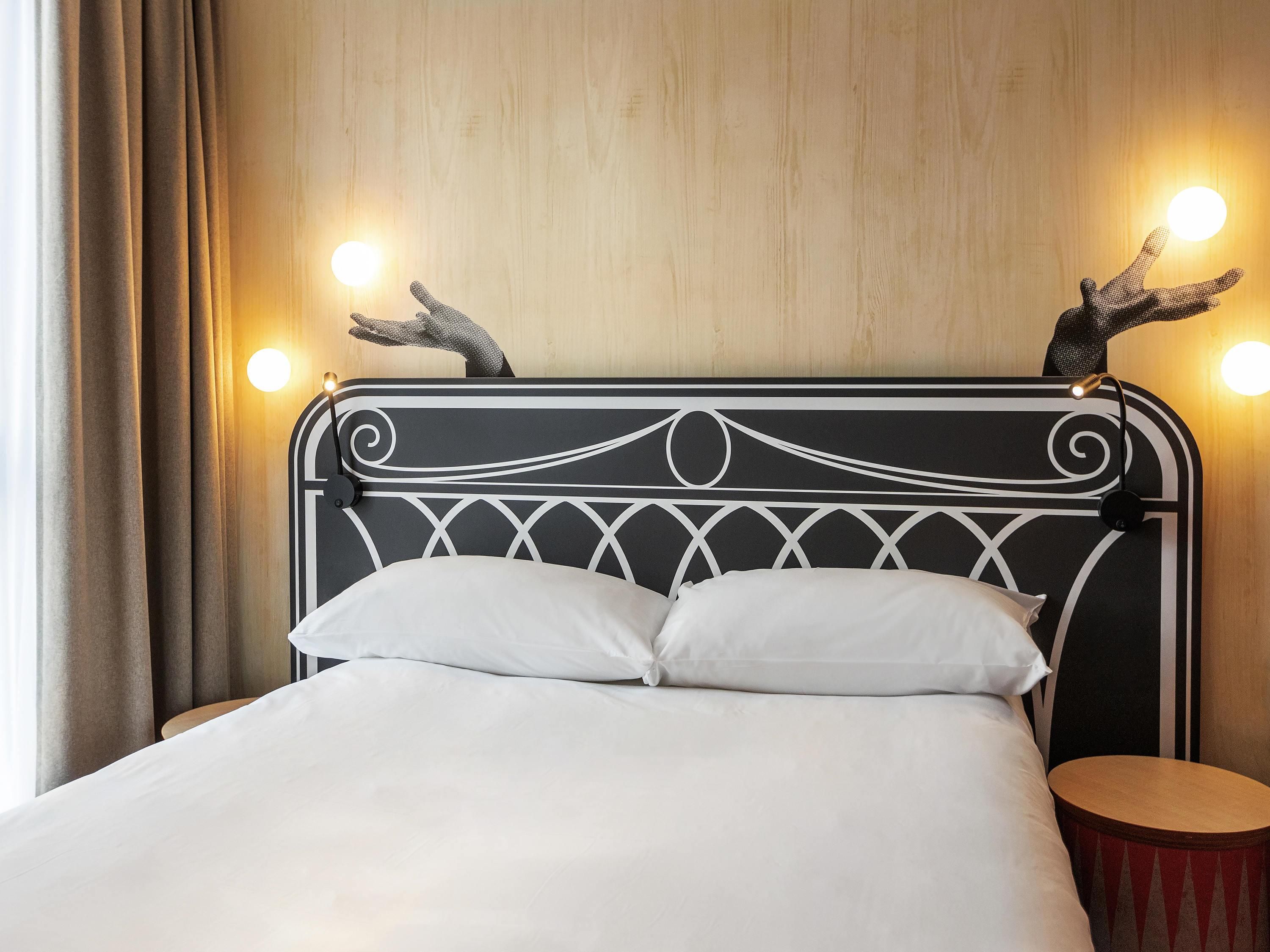 Una cama con cabecera de metal negro, dos almohadas blancas y un taburete de madera a su lado.