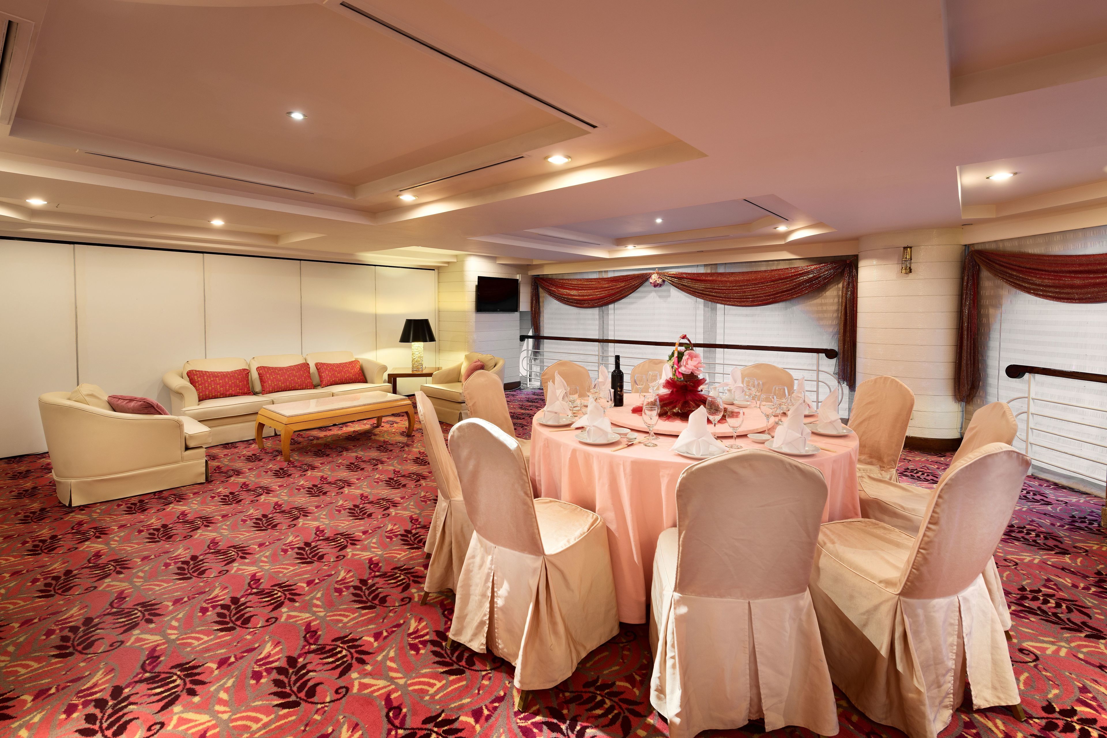 A room with a round table set for a meal, surrounded by chairs with light-colored covers. There is a sofa set with red cushions, a lamp, and a television on the wall.